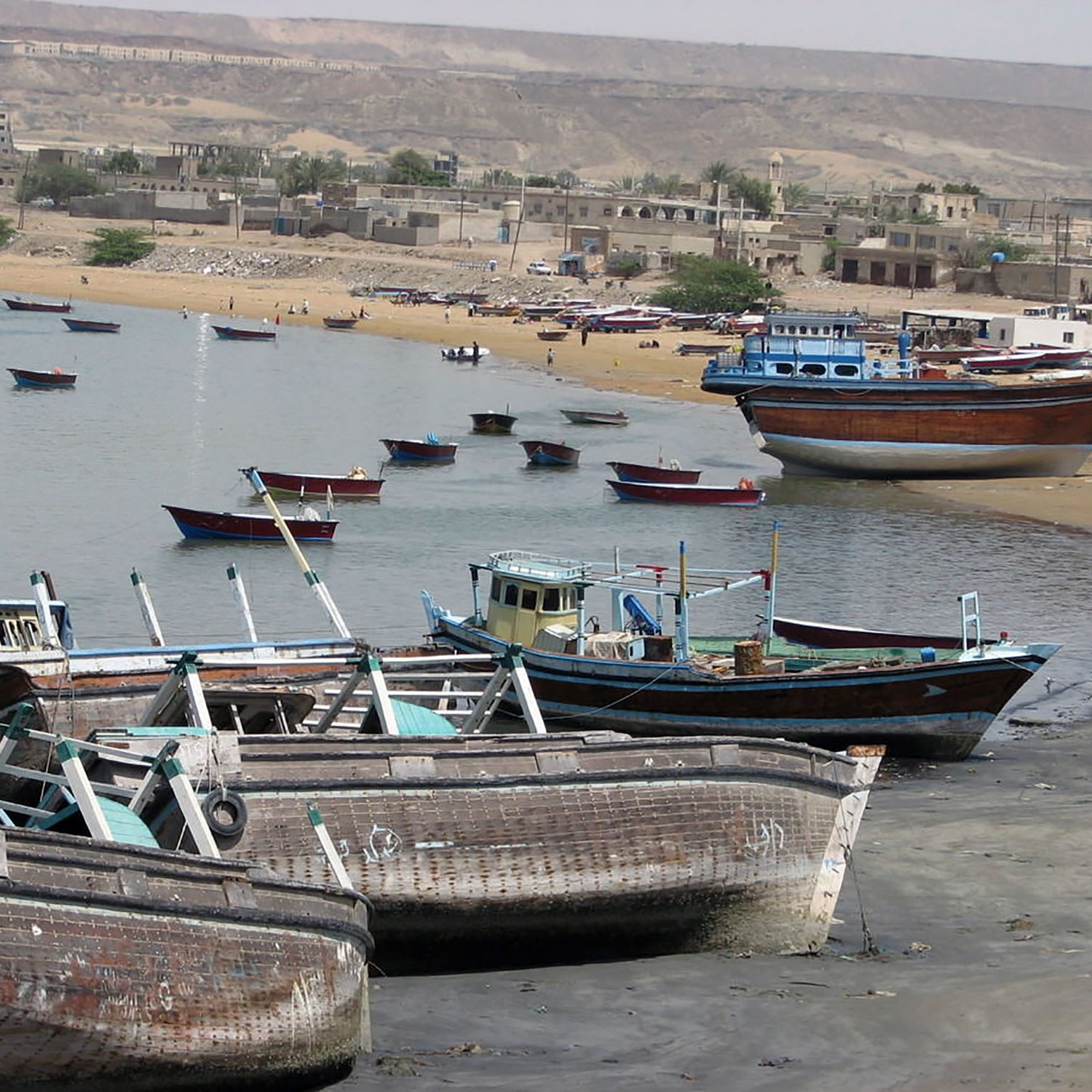 The image depicts a coastal scene with several fishing boats moored or resting on the shore. Some boats are partially submerged in shallow water, while others are docked along the beach. The background features sandy shores and low-lying buildings, with a mountainous area visible in the distance. The overall atmosphere suggests a tranquil seaside environment, typical of a fishing village.