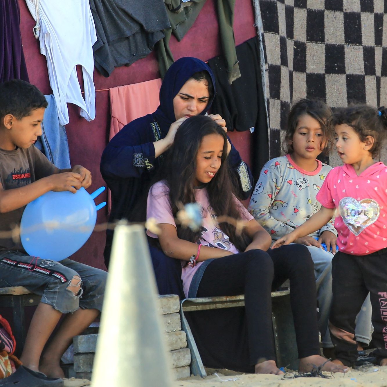 L'image montre un groupe d'enfants et une femme assis à l'extérieur, dans un environnement simple. Les enfants semblent interagir joyeusement, avec une petite fille qui parle à une autre. Une femme en vêtements traditionnels s'occupe des cheveux d'une jeune fille assise devant elle. En arrière-plan, on peut voir des vêtements accrochés et un sol en sable. L'ambiance est conviviale et émotive, évoquant des moments de partage et de complicité.
