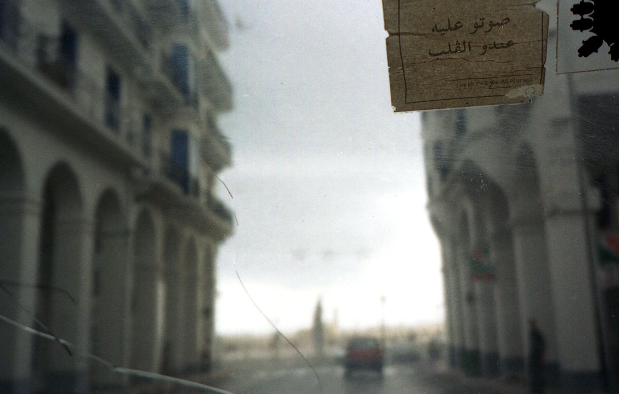 L'image présente une vue légèrement floue à travers un pare-brise. On peut apercevoir des bâtiments aux façades blanches avec des arches, typiques d'une architecture méditerranéenne. Sur le côté droit de l'image, un papier est collé sur le verre, avec des inscriptions en arabe. La scène semble se dérouler dans une rue calme, avec un véhicule au loin. L'ambiance générale est une combinaison de nostalgie et de tranquillité, suggérant une atmosphère de ville côtière. Le ciel est nuageux, ajoutant une touche de mélancolie à la composition.