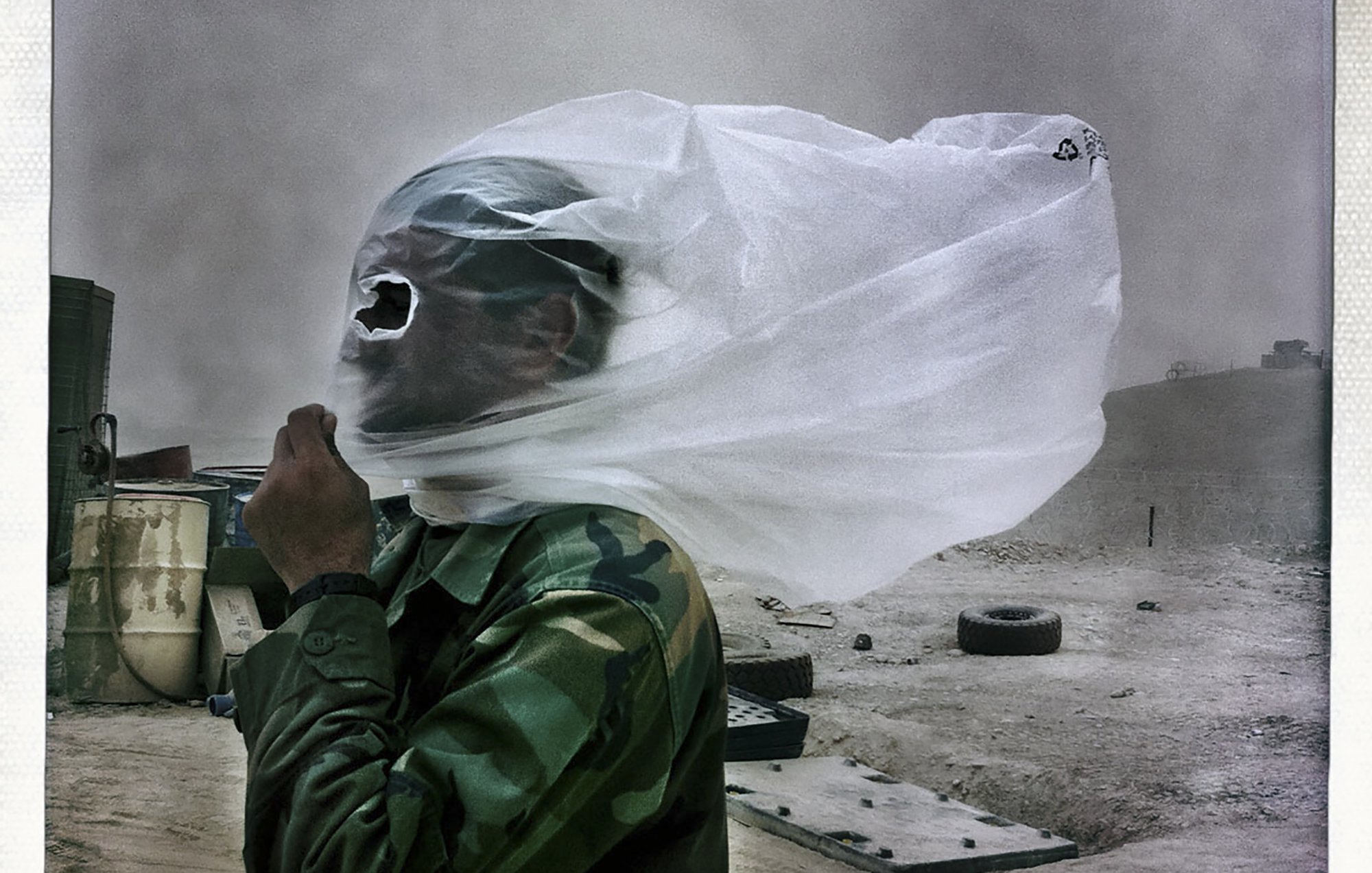La imagen muestra a un hombre vestido con un uniforme de camuflaje, protegiéndose de una tormenta de arena. Su rostro está cubierto con una tela transparente que parece ser una especie de máscara o protección. El entorno se ve polvoriento y desolado, con algunos objetos y neumáticos dispersos en el área. La escena transmite una sensación de dificultad y adversidad debido a las condiciones climáticas.