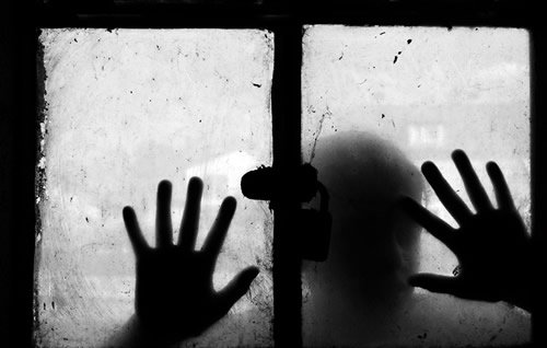 L'image montre une fenêtre sale avec un verre embué. Deux mains sont posées sur le verre, comme si quelqu'un tentait d'atteindre l'extérieur. La silhouette d'une personne est visible derrière le verre, ajoutant une atmosphère mystérieuse et mélancolique à la scène. L'utilisation du noir et blanc accentue le contraste et l'émotion de l'image, créant une sensation d'isolement et de confinement.