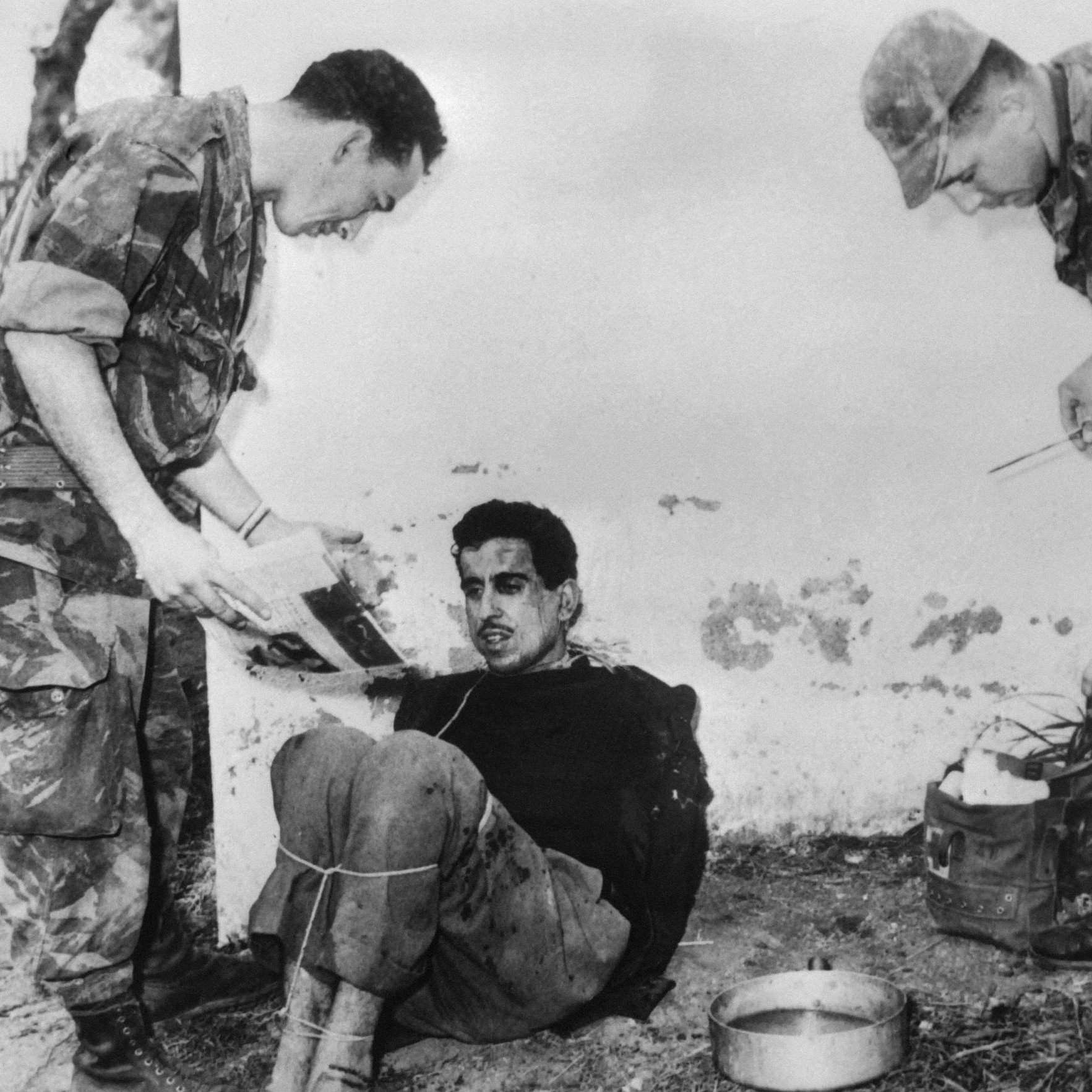 L'image montre un homme assis et attaché, visiblement en détresse, tandis que deux soldats, habillés en uniforme militaire, se tiennent près de lui. Un des soldats semble examiner un document ou une photographie, tandis que l'autre s'approche avec un équipement. L'environnement est sobre, et l'ambiance générale de la scène dégage un sentiment de tension.