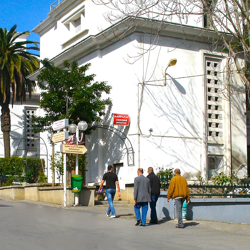 L'image montre une rue dégagée avec des bâtiments blancs et modernes de chaque côté. Des palmiers bordent le chemin, ajoutant une touche tropicale à l'environnement. On peut voir plusieurs personnes marchant le long de la rue, certaines en tenue traditionnelle. Le ciel est bleu et ensoleillé, ce qui donne une ambiance agréable et décontractée à la scène. Les façades des bâtiments présentent une architecture typique de la région, avec des détails subtils comme des lampadaires et des panneaux indiquant des directions.