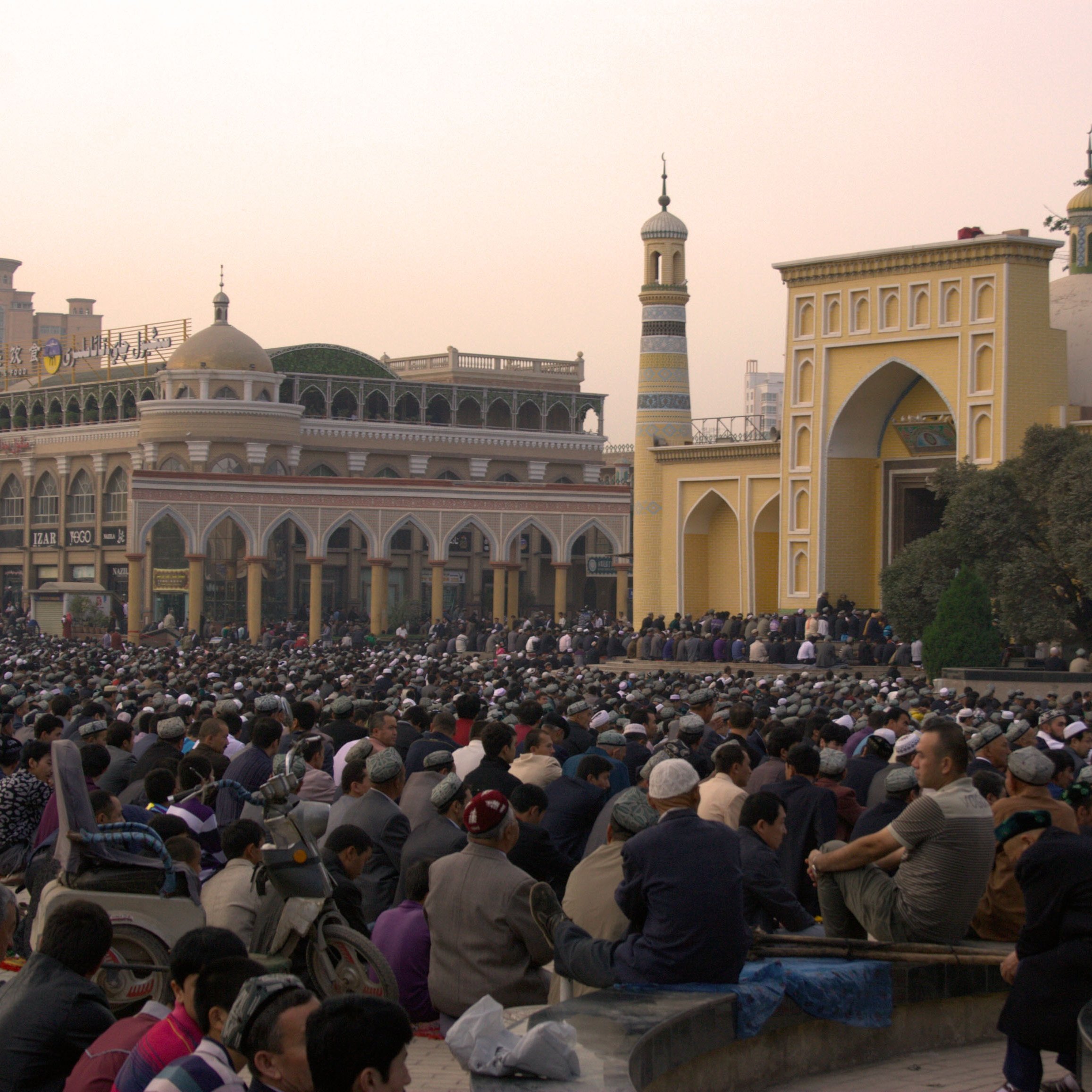 تش depicts الصورة تجمعًا كبيرًا من الناس، حيث يجلس عدد كبير من الأفراد في ساحة مفتوحة. في الخلفية، يمكن رؤية معمار تقليدي يتضمن مآذن وقباب، مما يشير إلى أنه قد يكون موقعًا دينيًا. السماء في حالتها النهارية تكتسي بألوان دافئة، مما يضفي جواً خاصاً على المشهد. يبدو أن الناس مجتمعون لأداء نشاط جماعي، ربما صلاة أو احتفال.