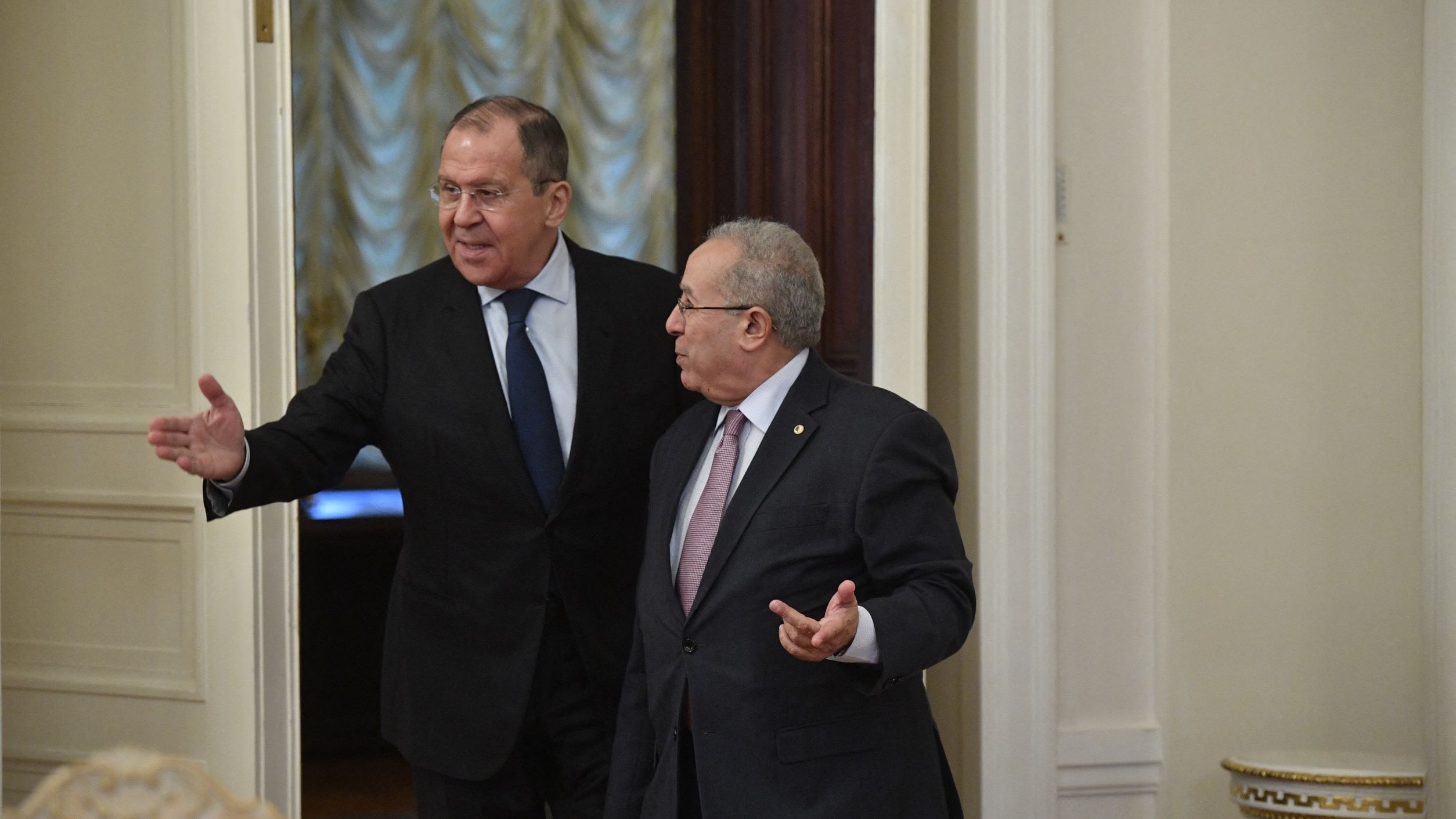 En la imagen se puede ver a dos hombres en un entorno formal, probablemente en una sala de reuniones o un despacho. Uno de ellos, a la izquierda, parece estar señalando o gesticulando con una mano, mientras que el otro, a la derecha, escucha y gira ligeramente su cuerpo hacia él. Ambos visten trajes formales y tienen expresiones serias. El ambiente parece elegante, con detalles en las paredes y una iluminación suave.