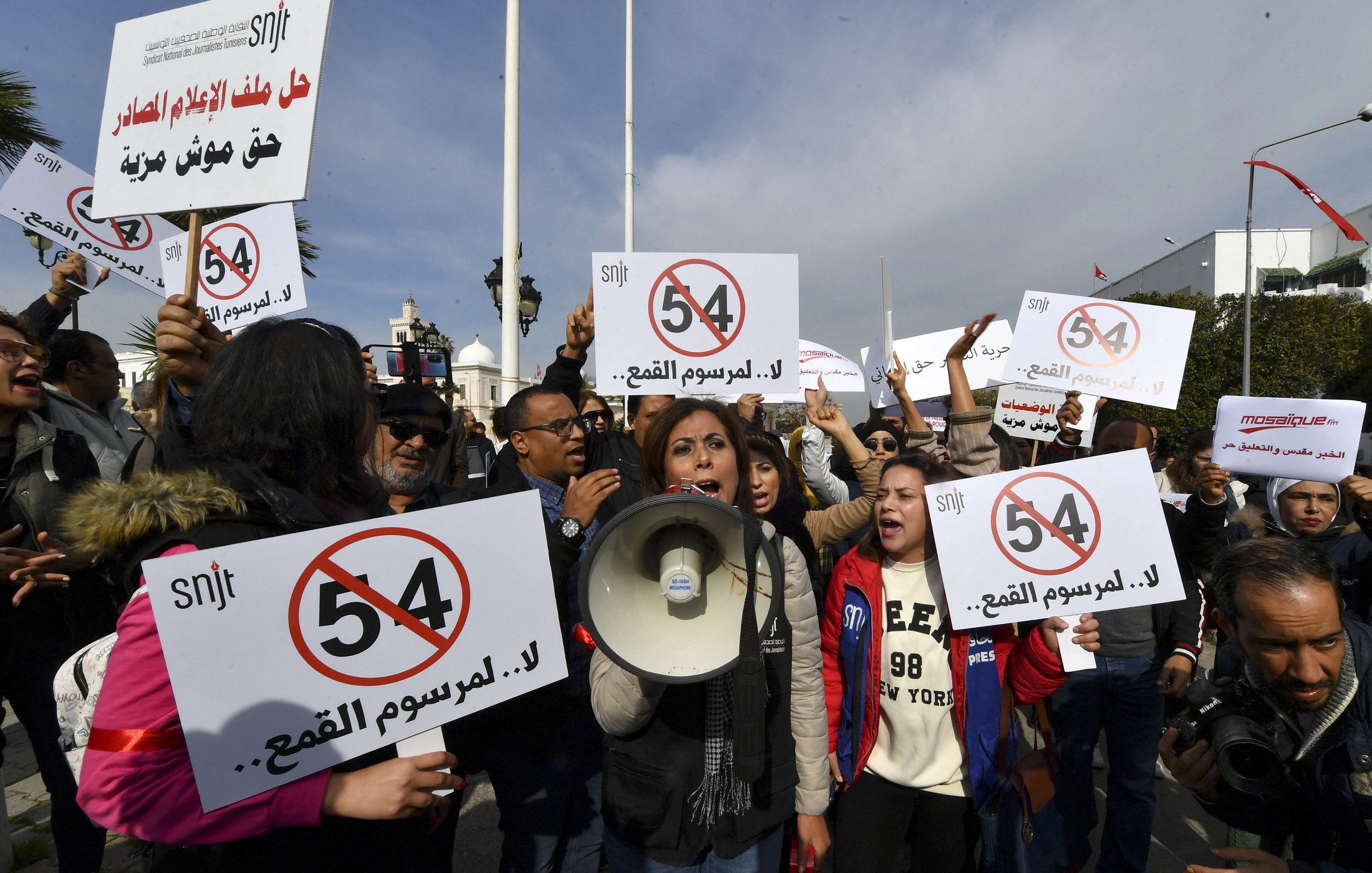 L'image montre une manifestation avec des groupes de personnes tenant des pancartes. Une femme au centre utilise un mégaphone pour s'exprimer. Les pancartes affichent des messages en arabe et en chiffres, notamment le chiffre "54", ainsi que des slogans contre "la censure". L'atmosphère semble énergique, avec des participants engagés dans la cause. Les drapeaux au fond ajoutent à l'ambiance de protestation.