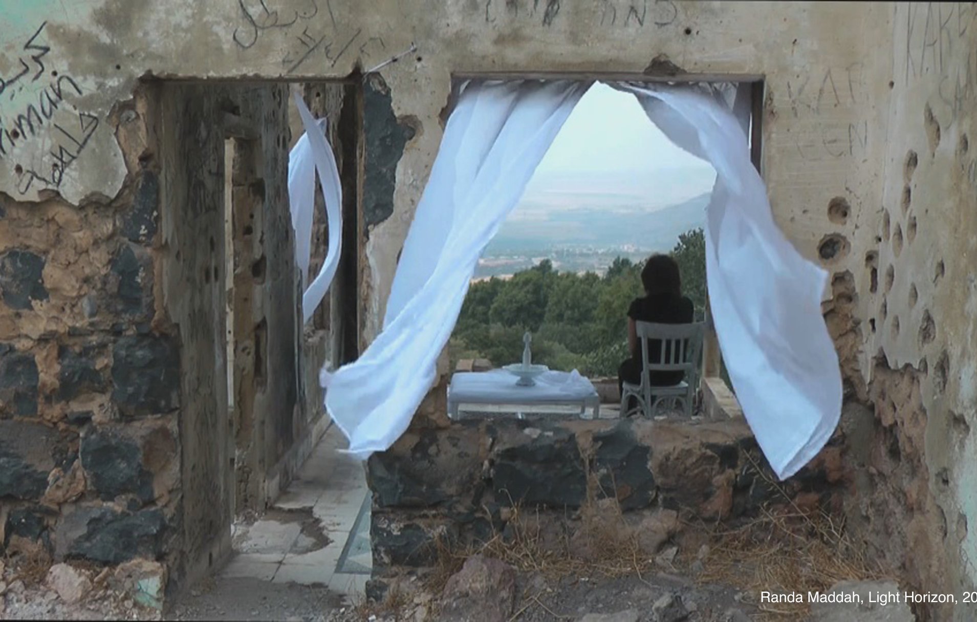 L'image montre une scène à l'intérieur d'un bâtiment partiellement ruiné. On peut voir une fenêtre encadrée par des rideaux blancs flottants. À travers cette fenêtre, un paysage verdoyant s'étend avec des collines au loin. À l'intérieur, une chaise blanche est placée près d'une petite table, créant une atmosphère de calme et de solitude. Les murs sont marqués par des graffitis, ajoutant une dimension de mémoire et d'histoire à l'espace.