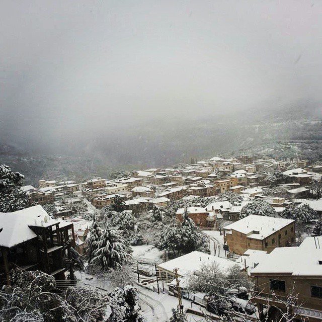 L'image montre un paysage hivernal où une petite ville est recouverte de neige. Les toits des maisons, en grande partie en bois, sont également blancs, créant une atmosphère paisible et froide. On peut apercevoir des arbres chargés de neige et des collines en arrière-plan, le tout enveloppé par une brume légère, ajoutant à la sérénité de la scène. La vue donne une impression de calme et d'isolement, typique des régions montagneuses en hiver.