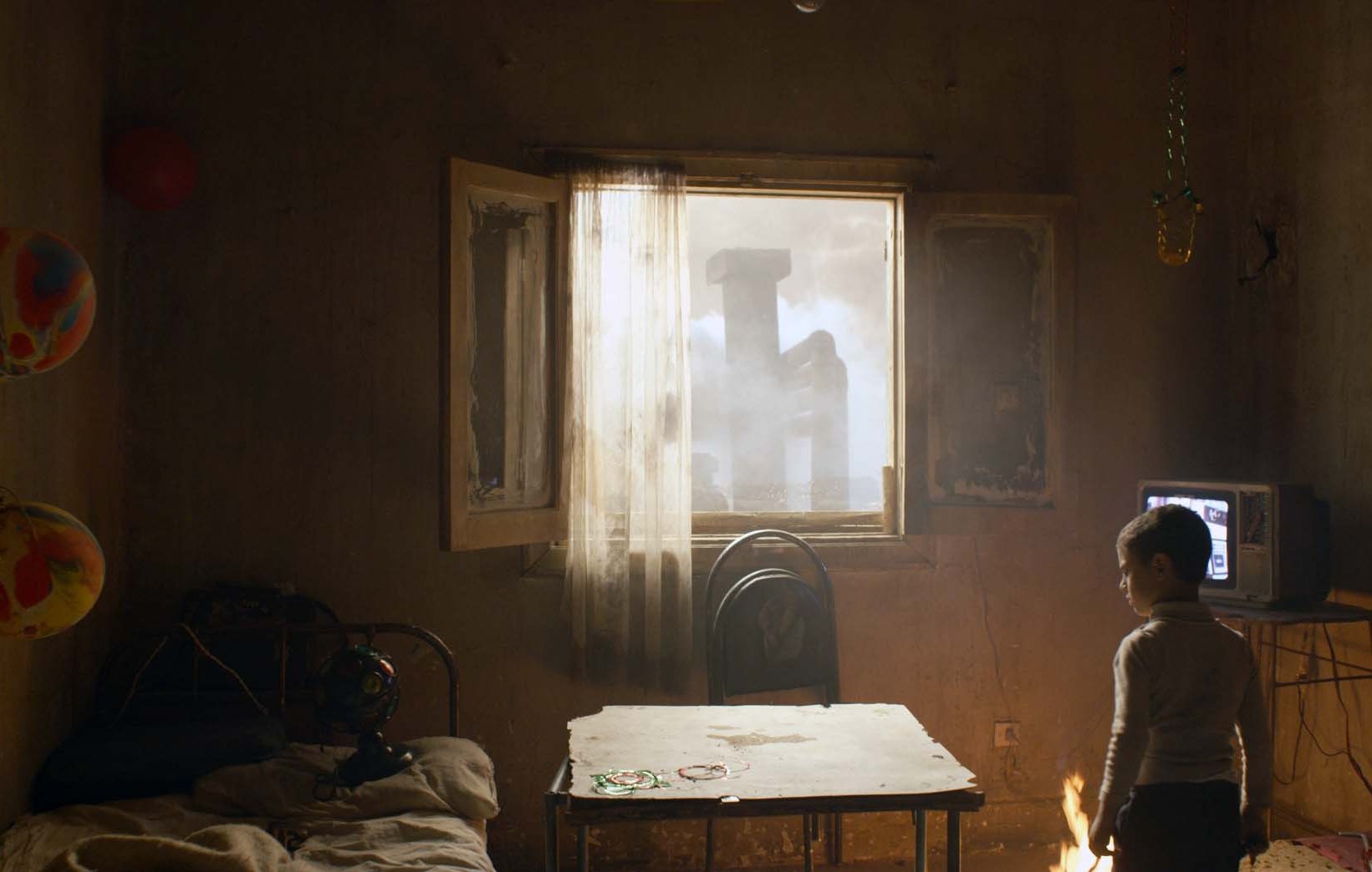 L'image montre une pièce sombre et légèrement en désordre, avec un enfant tourné vers une fenêtre ouverte. La lumière pénètre par cette fenêtre, révélant une scène extérieure brumeuse. À l'intérieur, on voit un lit, une table en bois et un vieux téléviseur. Des décorations festives, comme des ballons et des guirlandes, ajoutent une touche de couleur, bien que l'atmosphère générale semble mélancolique.
