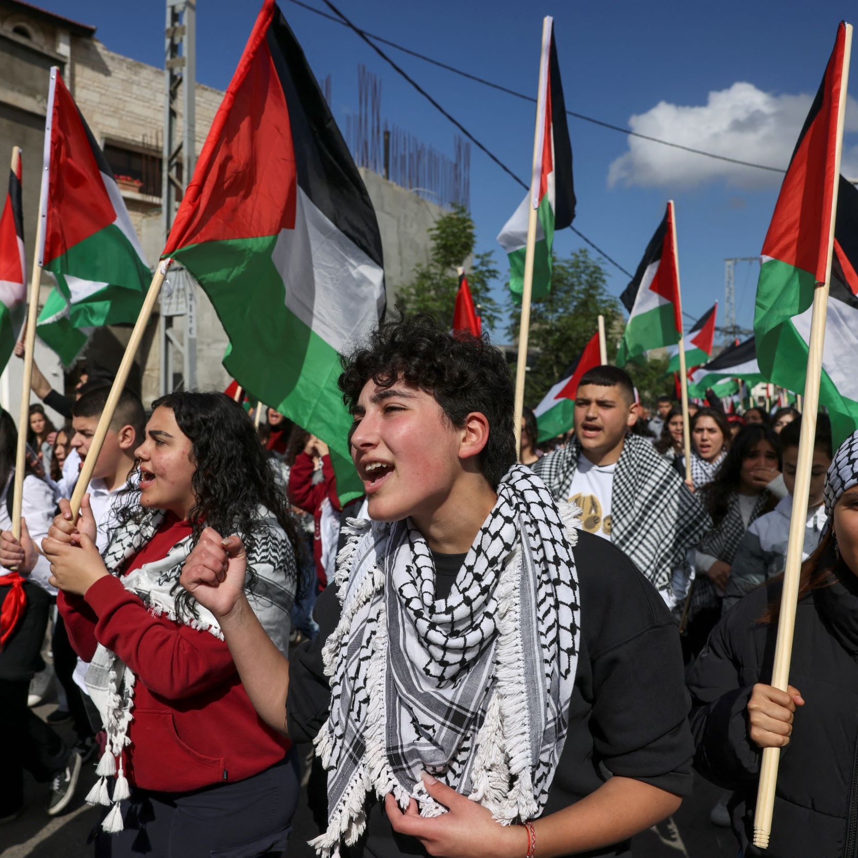 L'image montre une foule de manifestants, principalement des jeunes, participant à une marche ou une manifestation. Ils portent des drapeaux palestiniens aux couleurs rouge, noir, blanc et vert, et certains portent des keffiehs, des écharpes traditionnelles palestiniennes, souvent à motifs noirs et blancs. Les manifestants semblent passionnés et chantent, exprimant leur soutien à une cause. En arrière-plan, on aperçoit des bâtiments et un ciel partiellement nuageux.