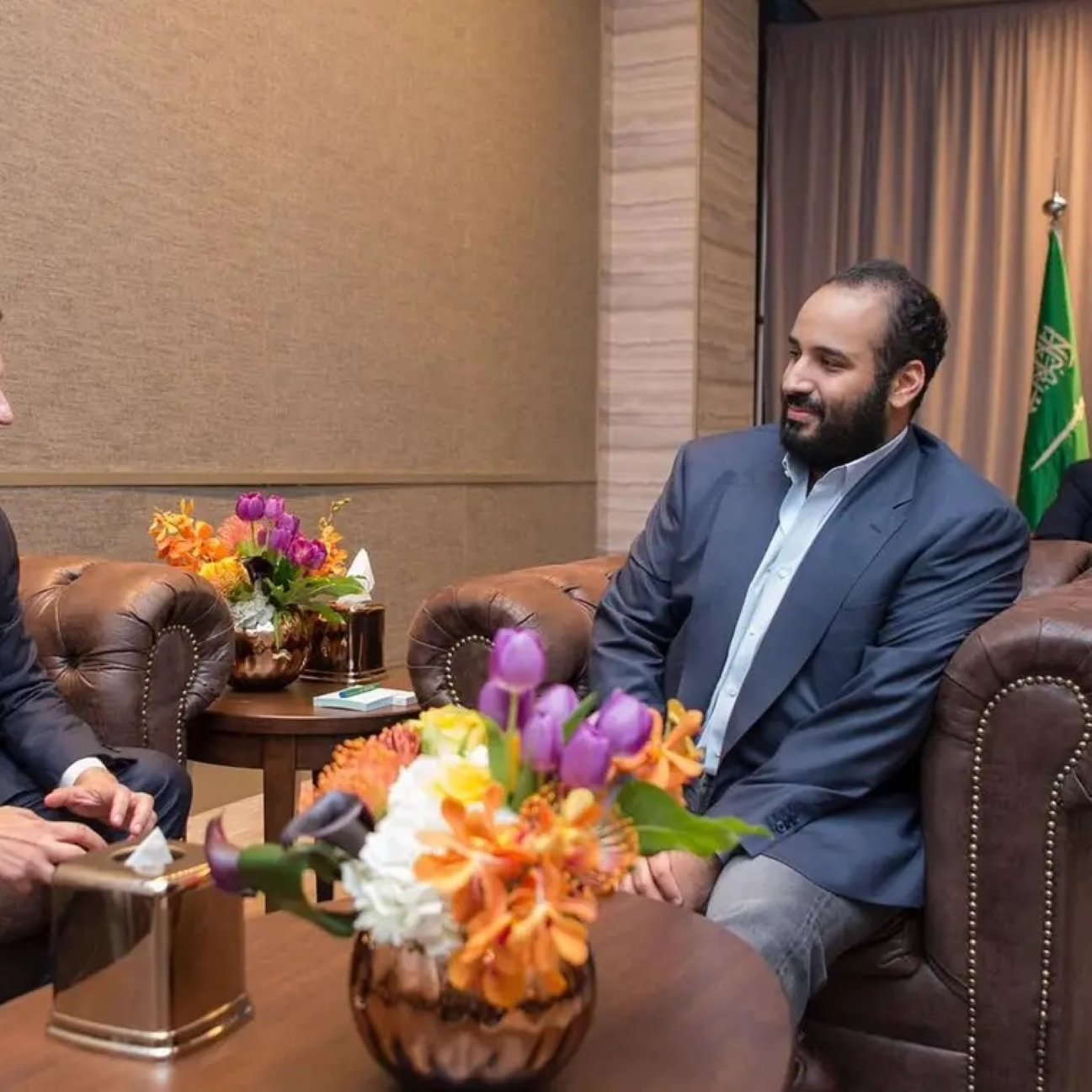 دو مرد در یک اتاق مرتب نشسته‌اند. یکی با کتی تیره و دیگری با پیراهن روشن. گل‌های رنگارنگ روی میز است.