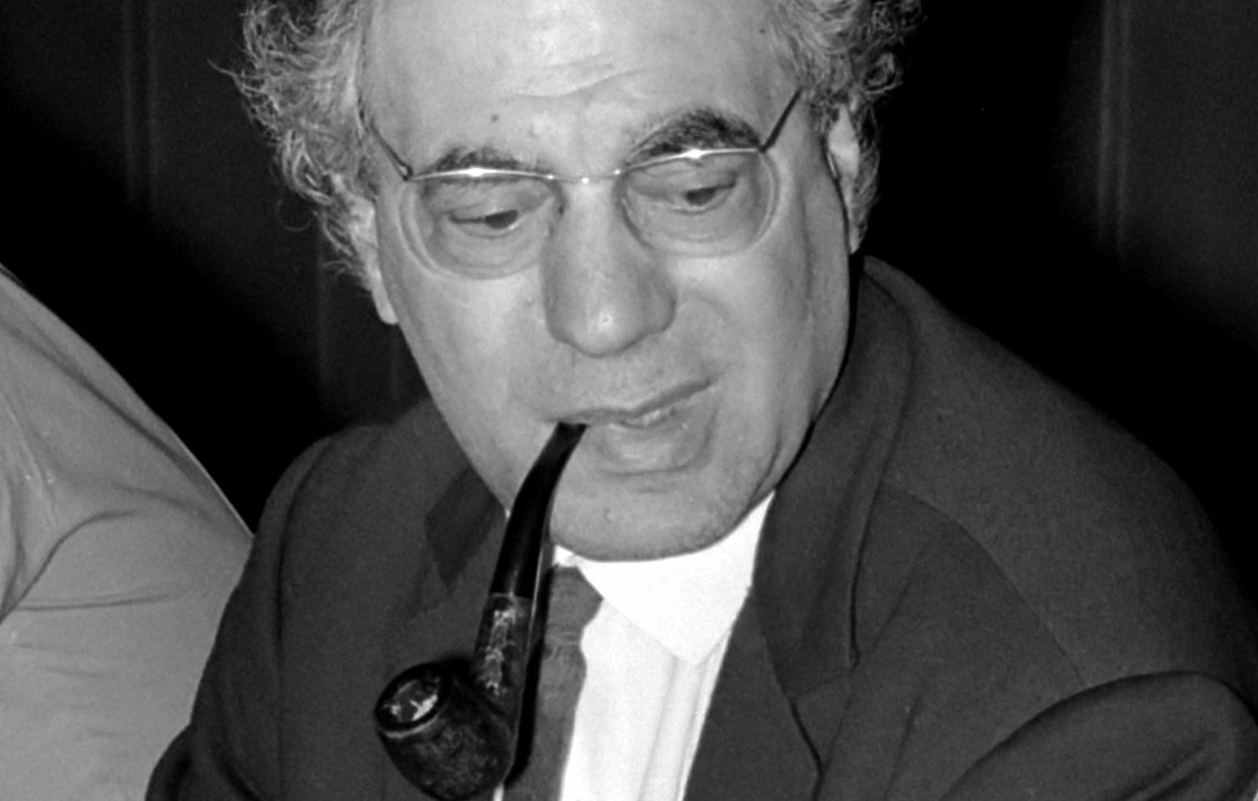 L'image montre un homme assis avec une pipe dans la bouche. Il porte une veste, une chemise blanche et une cravate. Ses cheveux sont grisonnants et légèrement bouclés. Il a des lunettes et semble concentré sur une conversation ou une discussion. En arrière-plan, on peut apercevoir d'autres personnes, mais elles ne sont pas clairement visibles. L'éclairage est modéré, ce qui donne une ambiance sérieuse à la scène.