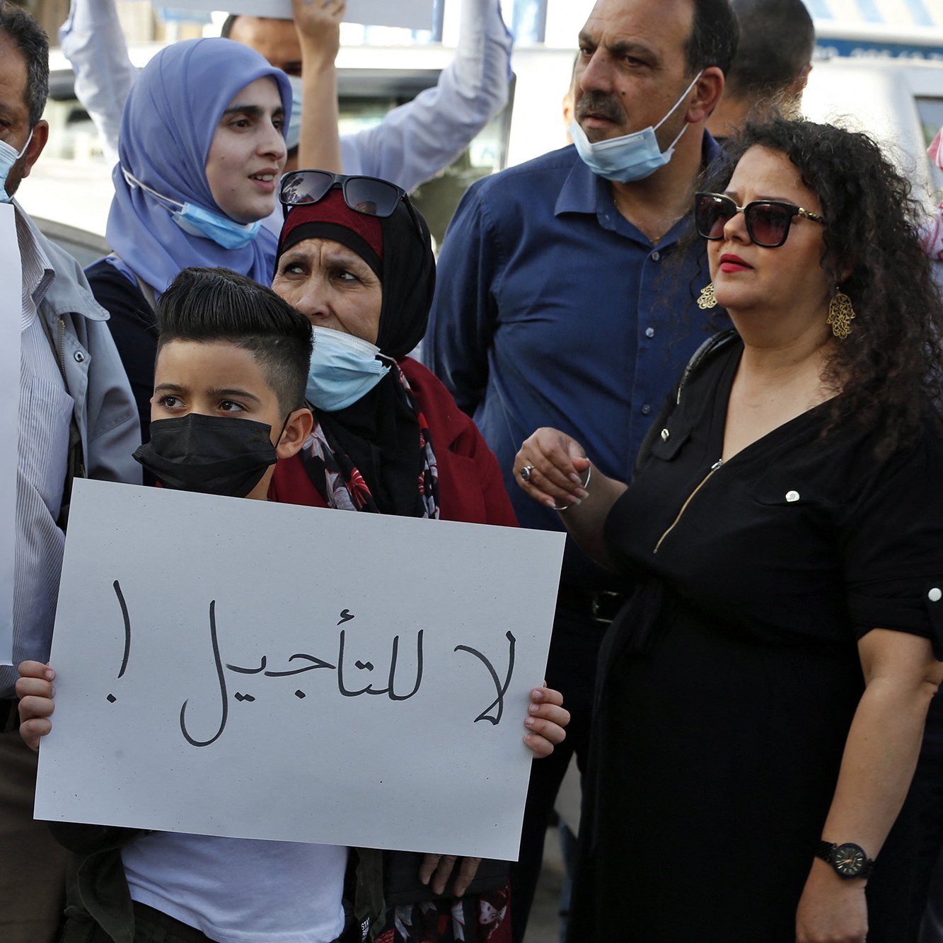 L'image montre un groupe de personnes rassemblées pour une manifestation. Parmi elles, il y a des adultes et des enfants, certains portant des masques. Une femme en noir se tient à droite, tandis qu'un jeune garçon au centre tient une pancarte avec un message en arabe. Les manifestants semblent exprimer des préoccupations ou des revendications, chacun affichant une expression déterminée. L'ambiance générale est celle d'une protestation pacifique.