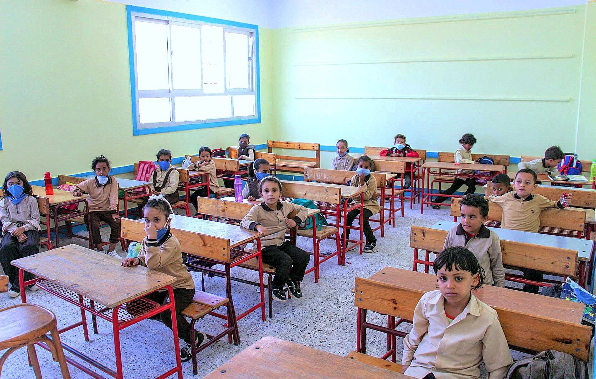 الصورة تُظهر فصلاً دراسياً يحتوي على عدد من الأطفال في مقاعدهم. الغرفة مضاءة جيداً بألوان فاتحة، الجدران مطلية بلون أخضر فاتح. الأطفال يرتدون زي المدرسة ويبدو عليهم الانتباه. بعضهم يرتدي كمامات. الطاولات والكراسي مصنوعة من الخشب. الغرفة منظمة ومرتبة وتحتوي على نوافذ كبيرة تسمح بدخول الضوء الطبيعي.