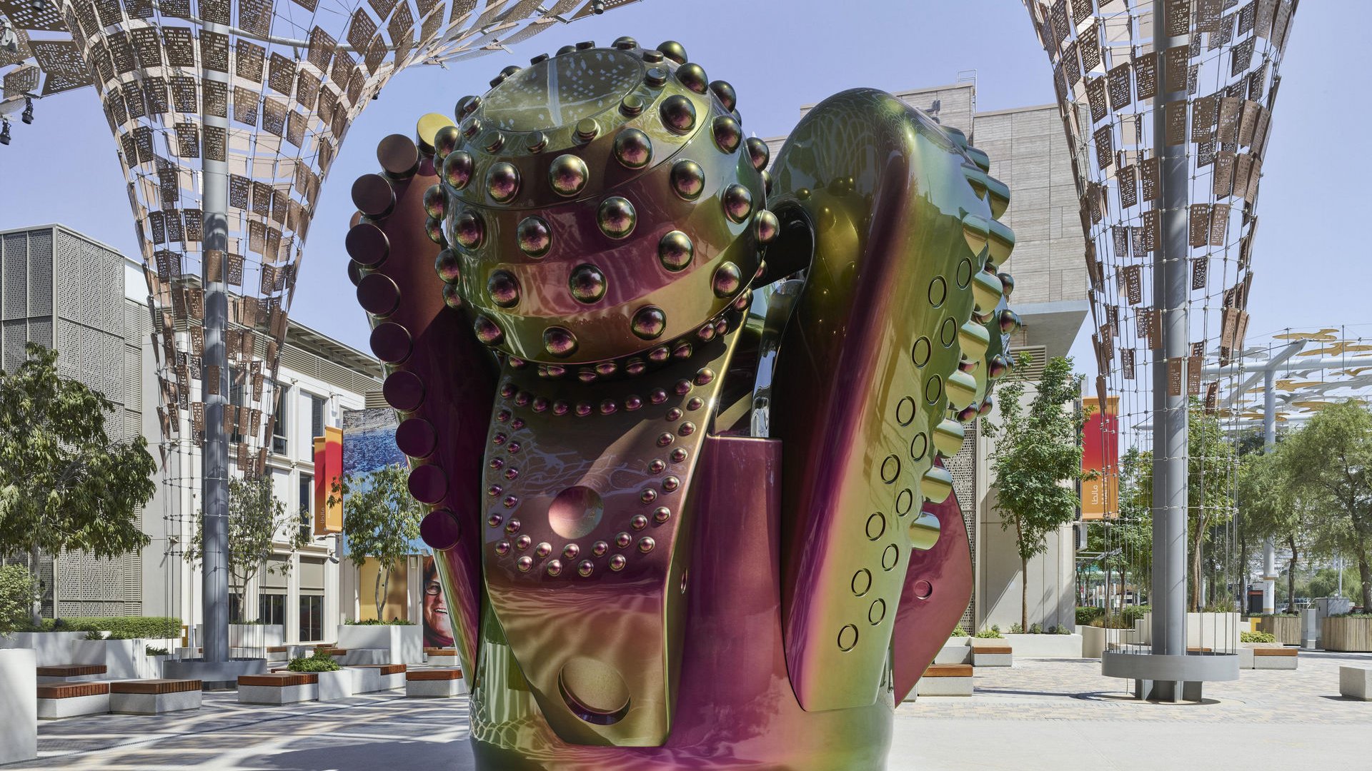 L'image présente une sculpture moderne au design futuriste, composée de formes organiques et géométriques. Les surfaces de la sculpture sont brillantes et colorées, avec des reflets irisés qui passent du vert au violet. Elle semble être placée sous une structure architecturale légère, qui offre de l'ombre et favorise une ambiance artistique. L'environnement autour de la sculpture est minimaliste, avec des arbres et quelques assises, ce qui met en valeur l'œuvre et crée une atmosphère apaisante.