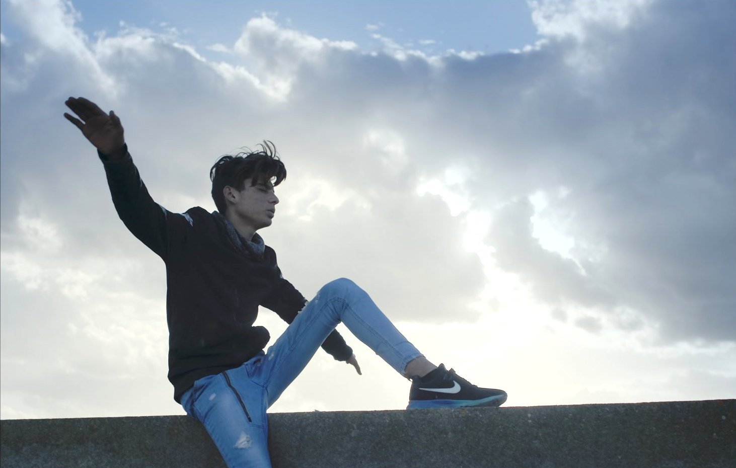 L'image montre un jeune homme assis sur un muret, avec une posture décontractée. Il porte un sweat noir et un jean bleu, avec des baskets noires. Ses cheveux sont légèrement ébouriffés par le vent, et il semble apprécier le moment en regardant vers le ciel, qui est nuageux avec quelques éclaircies. L'ambiance générale évoque une sensation de liberté et de fraîcheur.