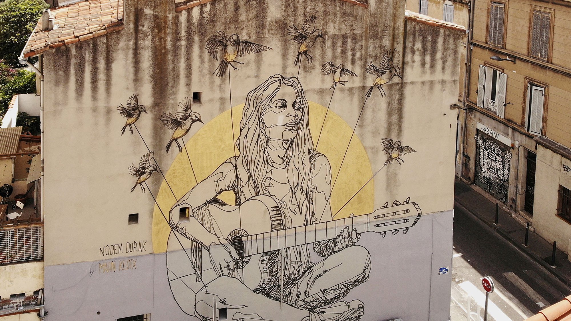 L'image montre une fresque murale colorée sur un bâtiment. Au centre, on voit une représentation d'une jeune femme assise, jouant de la guitare. Elle a de longs cheveux et un look décontracté. Autour d'elle, il y a plusieurs abeilles illustrées. En arrière-plan, on aperçoit des bâtiments typiques avec des fenêtres et des toits d'une ville. La fresque semble apporter une ambiance artistique et vivante à l'environnement urbain.