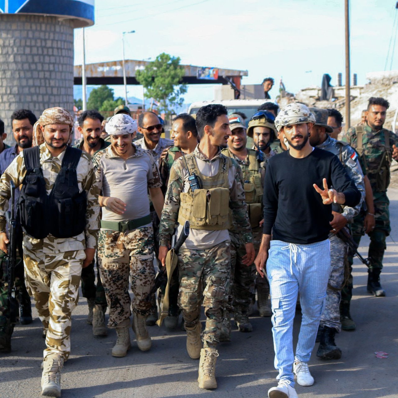 The image shows a group of soldiers and military personnel walking together in an outdoor setting. The soldiers are wearing various military uniforms and gear, including vests and helmets. Some are carrying weapons. In the foreground, a man in civilian clothes, showing a peace sign with his fingers, stands among them, smiling. The background suggests an urban area, with buildings and a road visible. The overall atmosphere appears to be one of camaraderie and unity.