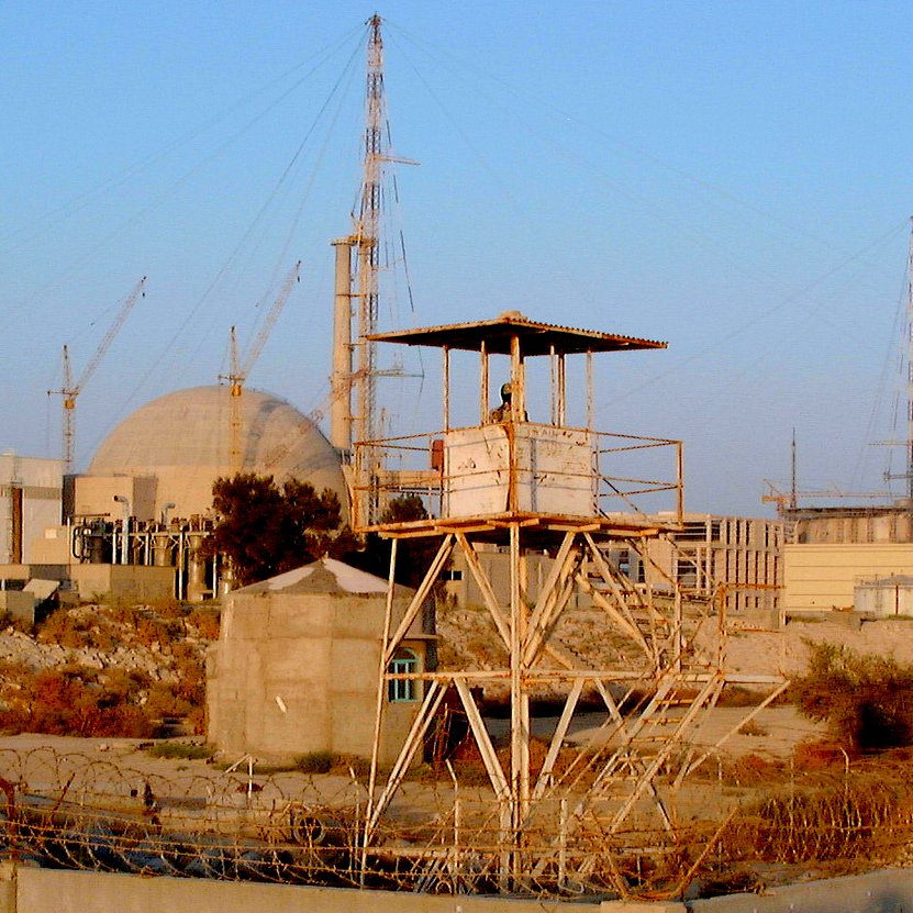 L'image présente un site industriel, probablement une centrale nucléaire, avec plusieurs structures. On peut voir une grande infrastructure circulaire au fond, entourée de diverses installations et grue. Au premier plan, il y a une tour de garde sur une plateforme surélevée, indiquant probablement un niveau de sécurité élevé dans cette zone. Des barbelés et des éléments de sécurité entourent l'endroit, suggérant une zone protégée. Le ciel est clair, ce qui donne une lumière naturelle à la scène.