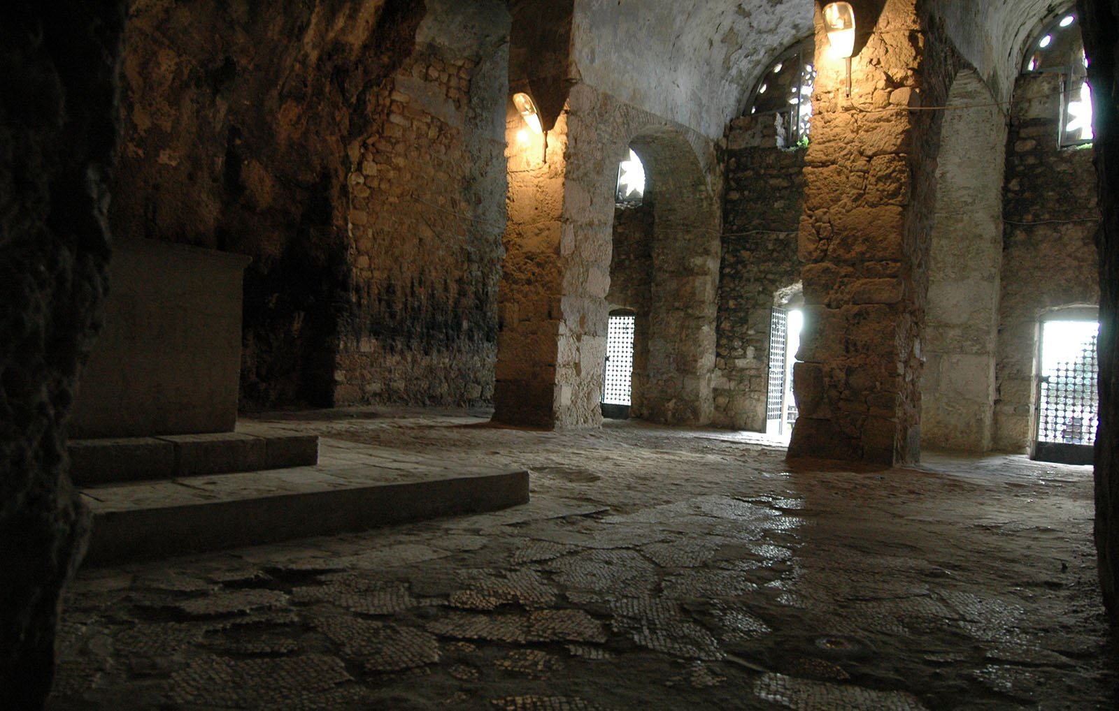 الصورة تُظهر مكانًا داخليًا يرجح أن يكون غرفة أو قبو بُني من الحجر. الجدران مكونة من حجارة كبيرة وغير منتظمة، وتعلوها بعض النوافذ المصفحة بالحديد مما يسمح بدخول الضوء الطبيعي. الأرضية مرصوفة بحجارة صغيرة، وقد تبدو رطبة بعض الشيء. الإضاءة في المكان خافتة، مما يضفي جوًا غامضًا على المكان. يمكن رؤية أجزاء من هيكل قديم يبدو أنه قديم جدًا.