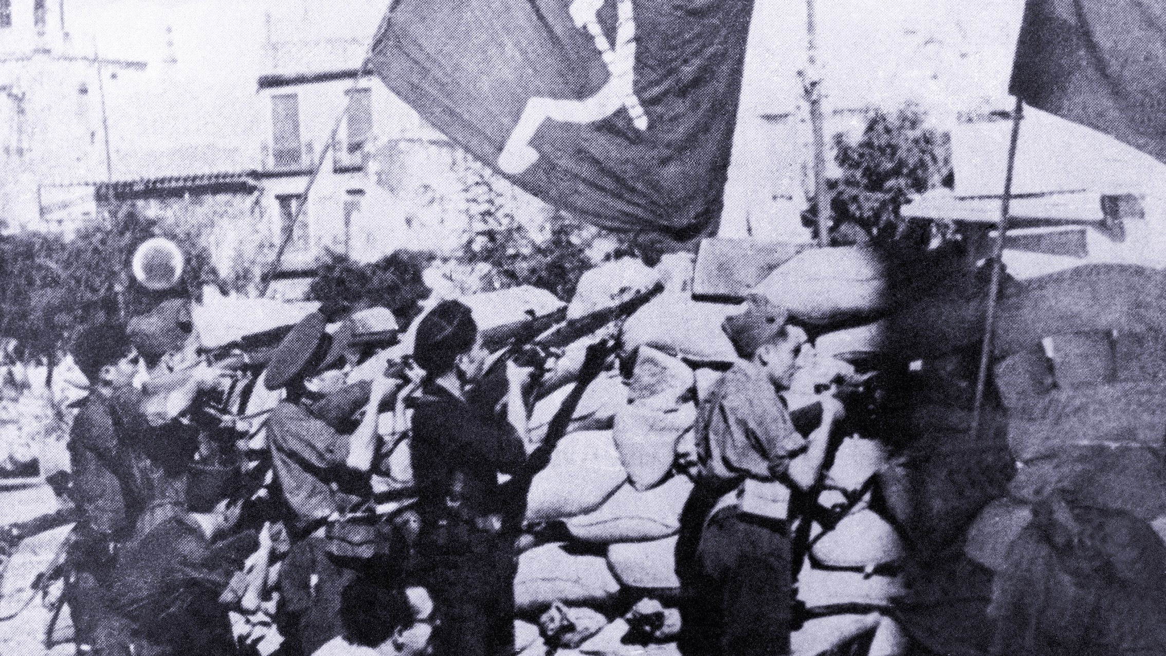 Cette image en noir et blanc montre un groupe de soldats ou de miliciens en position de tir, probablement lors d'un conflit. Ils se trouvent derrière un mur de sacs de sable, tout en tenant des fusils. En arrière-plan, des drapeaux avec un symbole communiste flottent, ce qui suggère un contexte politique et historique marqué par des idéologies de gauche. L'ambiance de la scène semble intense et sérieuse, indiquant un moment de bataille ou de lutte.