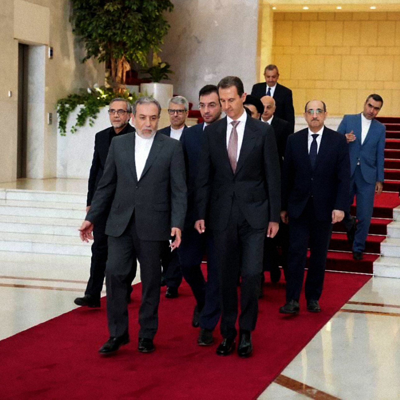 L'image montre un groupe de personnes marchant sur un tapis rouge dans un hall d'entrée élégant. À l'avant, deux hommes semblent en conversation, tandis que d'autres personnes les suivent, allant d'une manière formelle. L'environnement est spacieux avec des murs clairs et une décoration soignée, indiquant un lieu officiel ou diplomatique.