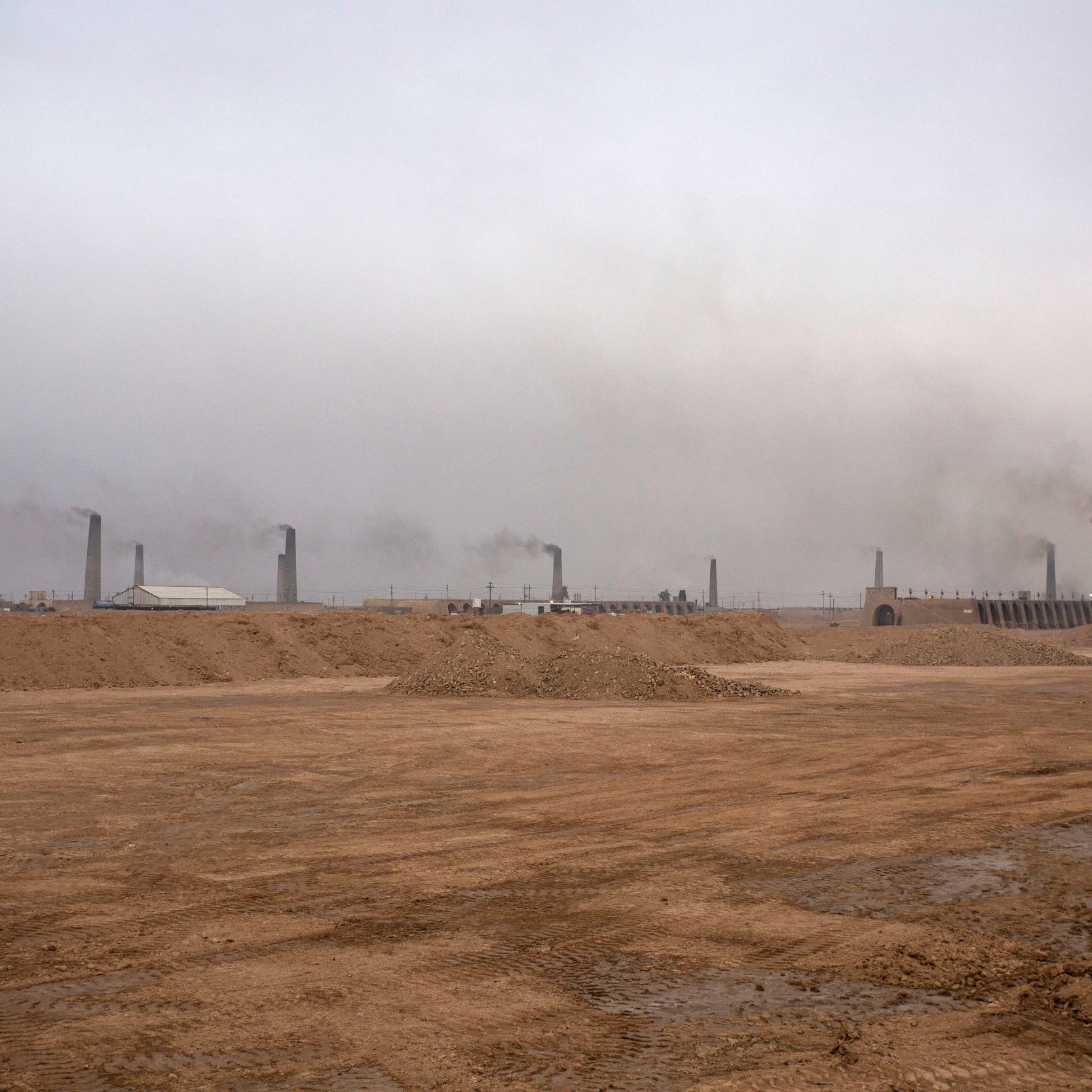L'image montre un paysage industriel avec un sol désertique et nu. À l'arrière-plan, de nombreux cheminées émettent de la fumée dans une atmosphère brumeuse et chargée. Le ciel est grisâtre, ce qui donne une impression de pollution et de dégradation environnementale. Le terrain est principalement constitué de sable ou de terre, et l'ensemble dégage une atmosphère morose et industrialisée.