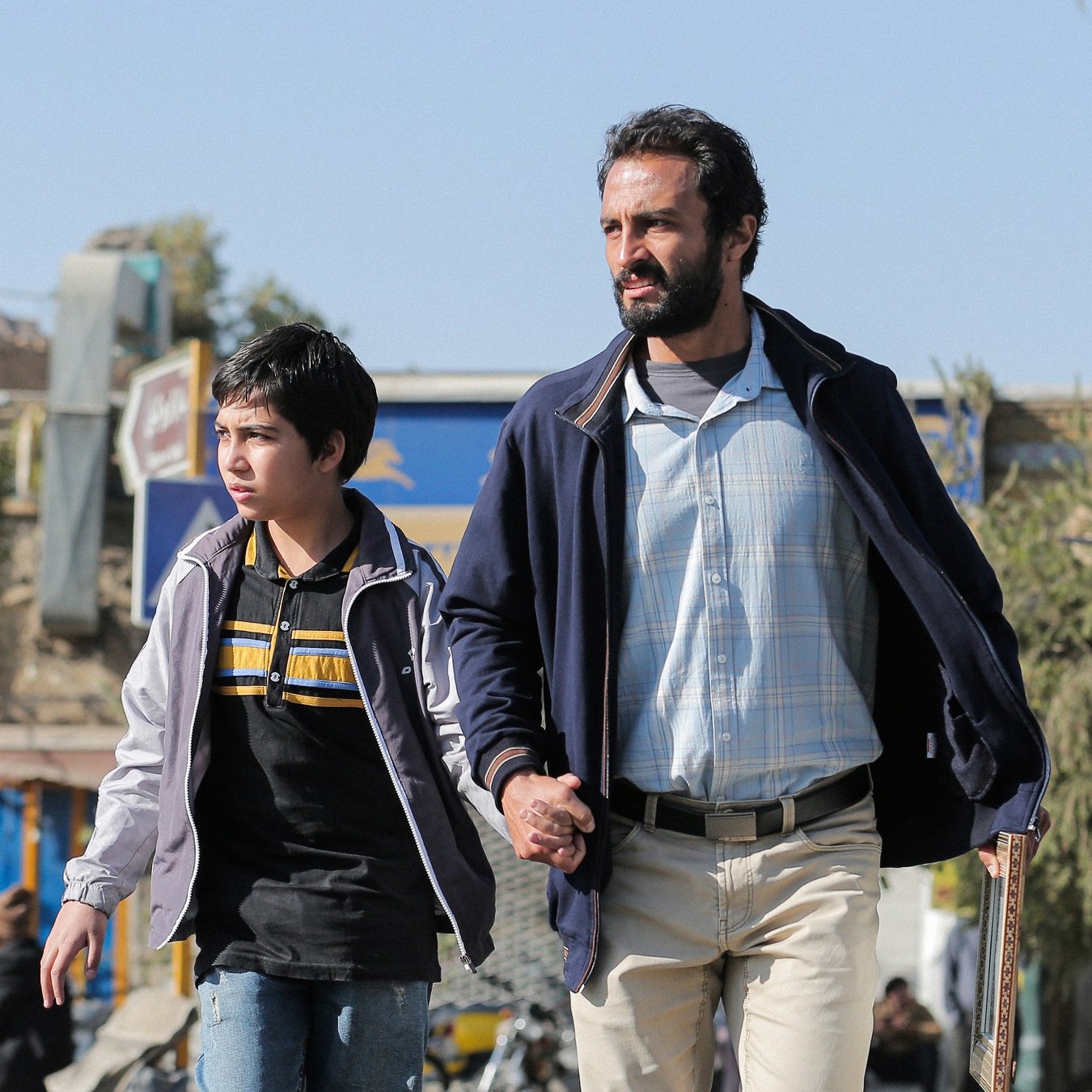L'image montre un homme et un garçon marchant ensemble dans une rue. L'homme, qui semble être un adulte, tient la main du garçon, qui est jeune et porte un haut à rayures. En arrière-plan, on aperçoit des bâtiments et des arbres, ce qui suggère un environnement urbain. L'éclairage est clair, indiquant probablement une belle journée. L'atmosphère semble à la fois tendre et protectrice, mettant en avant la relation entre les deux personnages.