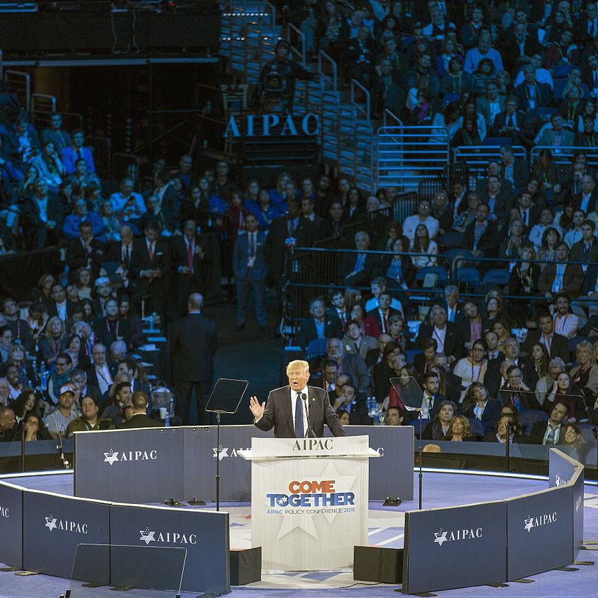 L'image montre un grand rassemblement dans une salle pleine de spectateurs. Au centre, une scène où un orateur, probablement un homme politique, s'exprime devant le public. Le logo "AIPAC" est visible sur des panneaux autour de la scène, indiquant que cet événement est lié à l'organisation pro-israélienne. Les participants semblent attentifs, et l'ambiance générale évoque un moment solennel et engagé. Les couleurs de l'éclairage sont principalement bleu et violet.