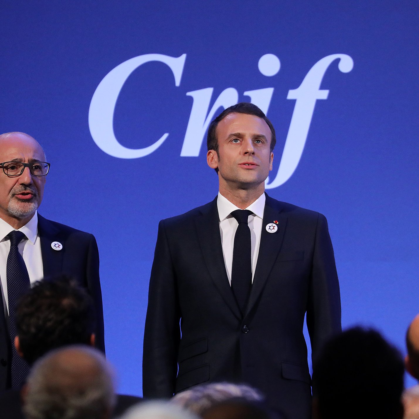 The image shows two men standing side by side in front of a blue backdrop with the word "Crif" displayed prominently. One man is dressed in a black suit and tie, appearing to be delivering a speech or presentation. The other man, also in a formal suit, stands next to him, possibly in a supportive or attentive role. The setting suggests a formal event, likely related to the organization represented by "Crif."