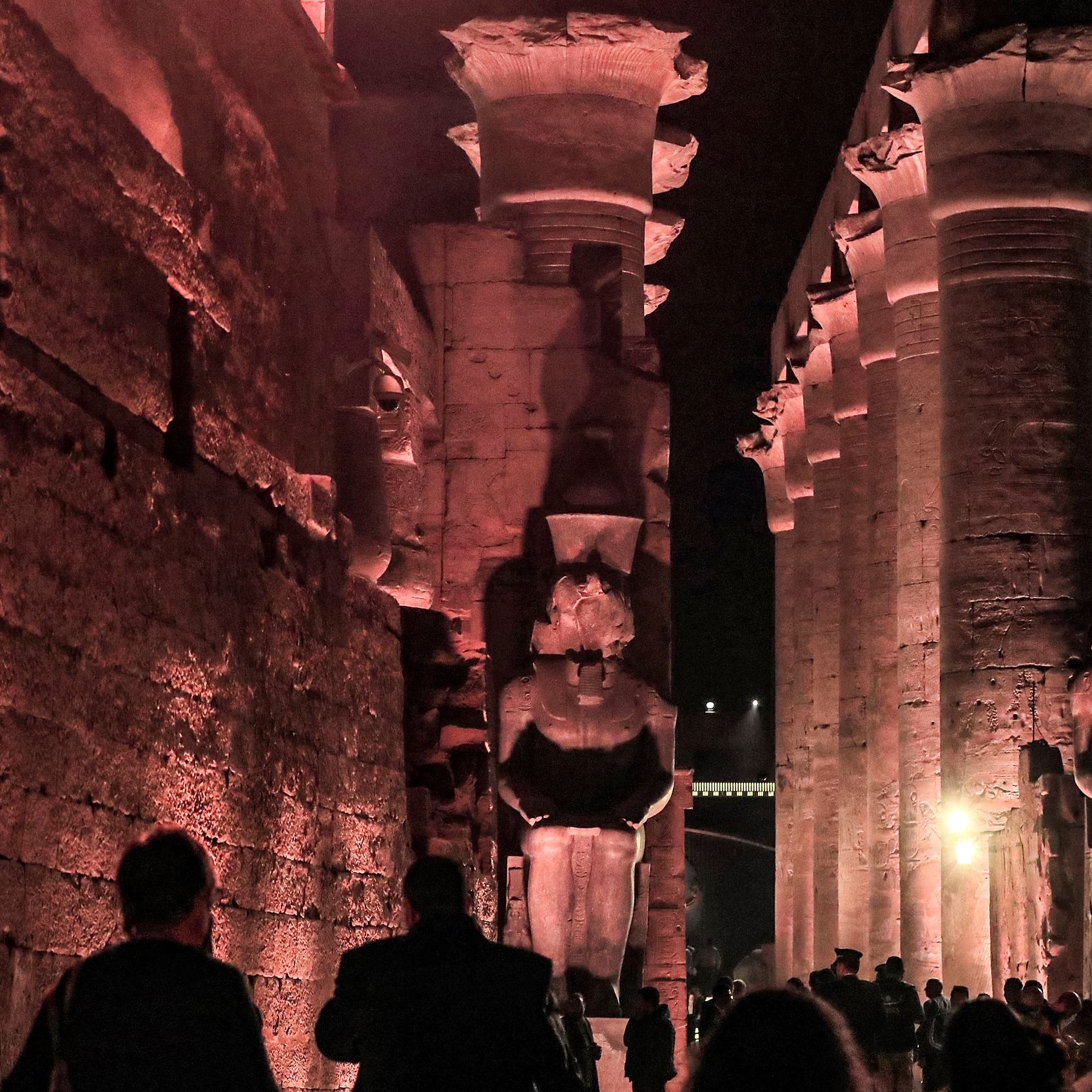 الصورة تظهر معالم أثرية قديمة، حيث يمكن رؤية أعمدة ضخمة ومعمار تاريخي فاخر. الإضاءة الليلية تضفي جوًا سحريًا على المكان، مع تماثيل كبيرة تشكل جزءًا من التصميم المعماري. يبدو أن هناك زوارًا يستكشفون هذا الموقع التاريخي، مما يدل على شهرة وأهمية هذا المكان السياحي.