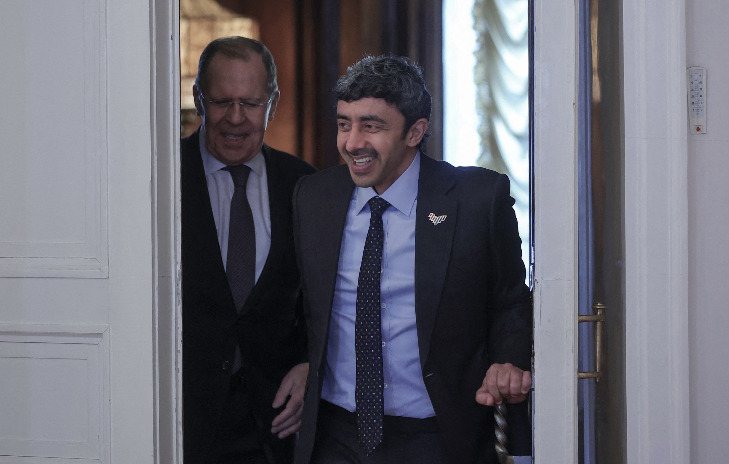 L'image montre deux hommes entrant dans une pièce. L'un d'eux sourit et semble de bonne humeur, tandis que l'autre a une expression plus neutre. Ils portent des costumes assortis et se trouvent dans un environnement élégant avec des éléments de décoration classique. La lumière naturelle entre par une fenêtre, ajoutant une ambiance chaleureuse à la scène.