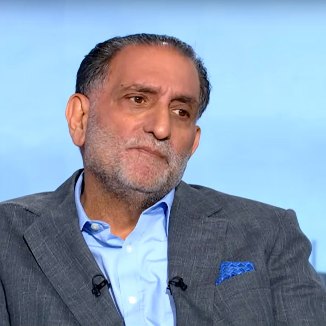 A middle-aged man in a suit with a serious expression, seated against a blurred background.