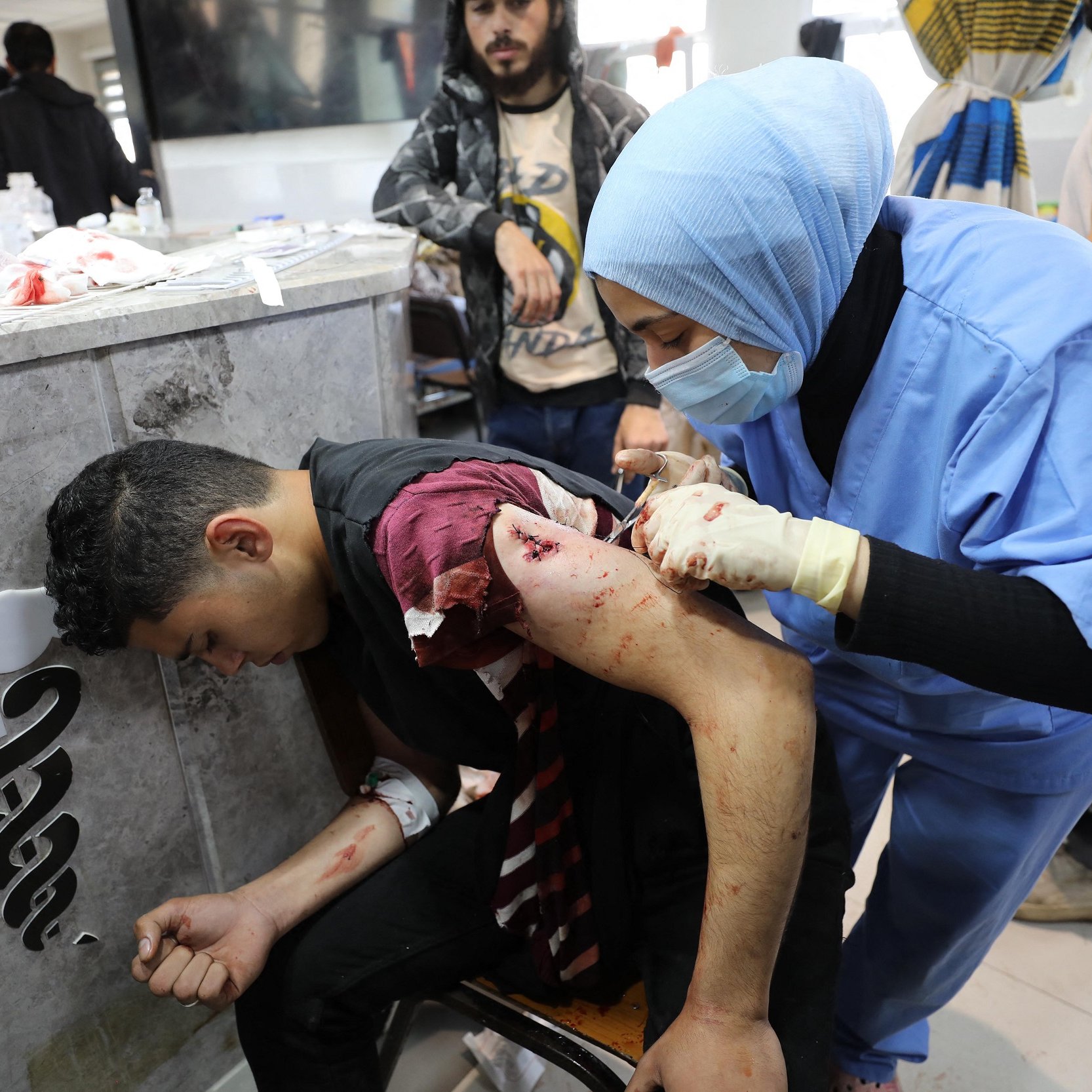 تصویر یک صحنه پزشکی را نشان می‌دهد. در این تصویر، یک نوجوان یا جوان با زخم‌های جدی بر روی بازوی خود نشسته است و یک پرستار یا پزشک در حال درمان اوست. او با لباس‌های پزشکی آبی و دستکش، مشغول پانسمان و رسیدگی به زخم‌های بیمار است. حال عمومی بیمار نشان‌دهنده‌ی وضعیت اضطراری اوست و محیط اطراف نیز شلوغ به نظر می‌رسد.