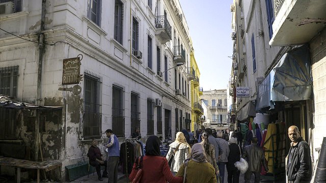 L'image montre une ruelle animée avec des bâtiments à l'architecture typique, dont certains sont peints en blanc et en jaune. On peut voir des gens marchant dans la rue, dont des femmes portant des habits traditionnels. À gauche, il y a un marché ou des stands avec des marchandises. L'ambiance semble vivante et typique d'un quartier urbain. La lumière du soleil éclaire la scène, ajoutant une chaleur à l'atmosphère.