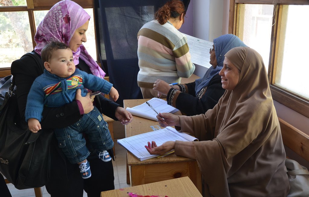 L'image montre une scène dans un environnement communautaire. On peut voir une femme portant un foulard qui tient un bébé dans ses bras, s'approchant d'une table où une autre femme, également couverte d'un foulard, est assise et prend des notes. À l'arrière-plan, d'autres femmes semblent occupées à diverses activités. L'ambiance semble être celle d'un événement organisé, possiblement une inscription ou une réunion. Les couleurs et les vêtements des femmes reflètent une culture riche et diversifiée.