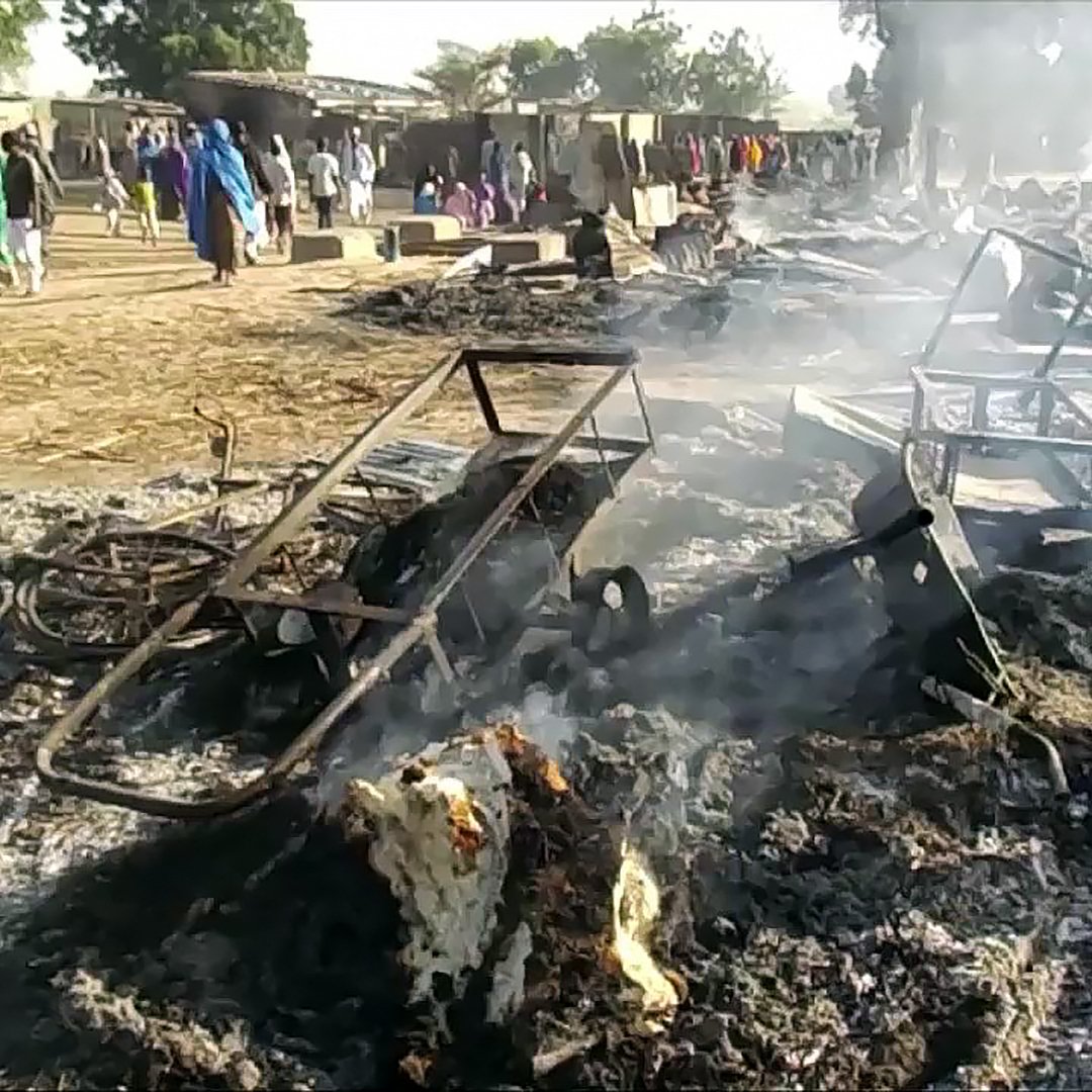 The image depicts a scene of destruction and aftermath, showing charred remains of structures and debris on the ground, with smoke rising. Various burned-out materials, such as metal frames and remnants of possibly furniture, are scattered across the area. In the background, a group of people can be seen, moving in what appears to be a community area, amidst the devastation. The setting suggests a recent fire or significant damage event, with the landscape appearing ashen and scattered with remnants of what once existed.