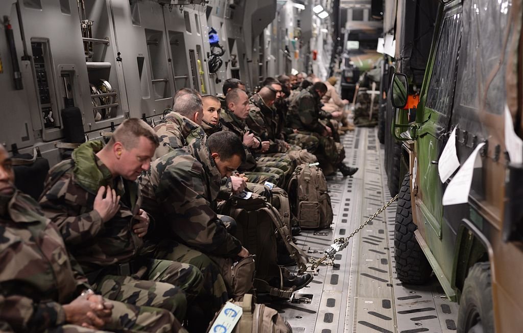 در این تصویر، گروهی از سربازان در داخل یک هواپیما یا وسیله نقلیه نظامی نشسته‌اند. سربازان در لباس‌های camouflage (رنگ‌های نظامی) دیده می‌شوند و به سمت جلو نشسته‌اند، در حالی که هر کدام یک کوله‌پشتی دارند. فضای داخلی وسیله نقلیه به نظر مدرن می‌رسد و تجهیزات نظامی در اطراف آن قرار دارد. برخی از سربازان به نظر می‌رسد در حال آماده‌باش بوده و توجه‌شان به جلو معطوف است.