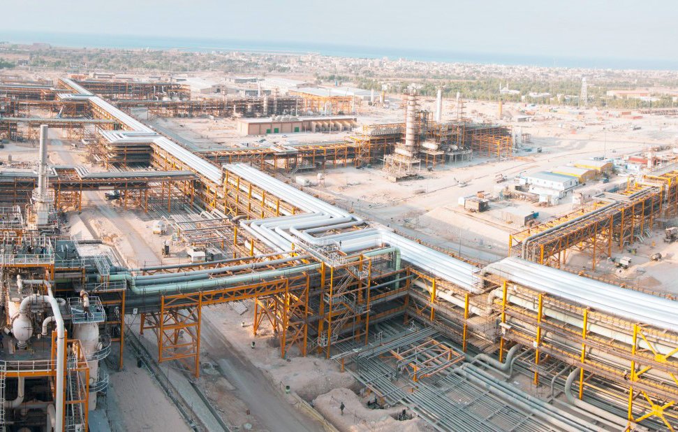 این تصویر یک مجتمع صنعتی بزرگ را نشان می‌دهد که شامل ساختارهای فلزی و لوله‌های گسترده است. در پس‌زمینه، تعدادی سازه بزرگ و تاسیسات به چشم می‌خورد. زمین اطراف به شکل خاکی و خشن است و در دوردست،‌ نوار آبی دریا نمایان است. این مکان به نظر می‌رسد که در حال فعالیت‌های صنعتی مانند پالایش نفت یا گاز باشد. فضا به طور کلی شلوغ و پر از تجهیزات و سازه‌ها است.