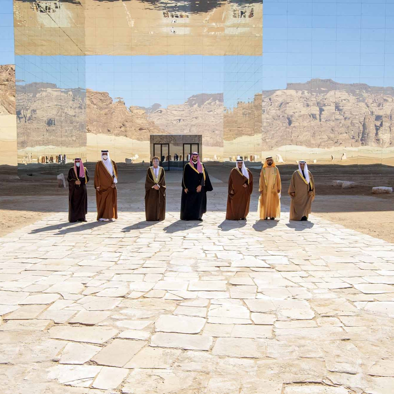 تظهر في الصورة مجموعة من الأشخاص يرتدون الزي التقليدي، ويقفوا على طريق مرصوف بالحجارة في منطقة صحراوية. خلفهم توجد جدران عاكسة تعكس المنظر الجبلي المحيط، مما يعطي شعوراً بالتواصل بين البشر والطبيعة. السماء زرقاء وصافية، مما يضيف جمالاً على المشهد.