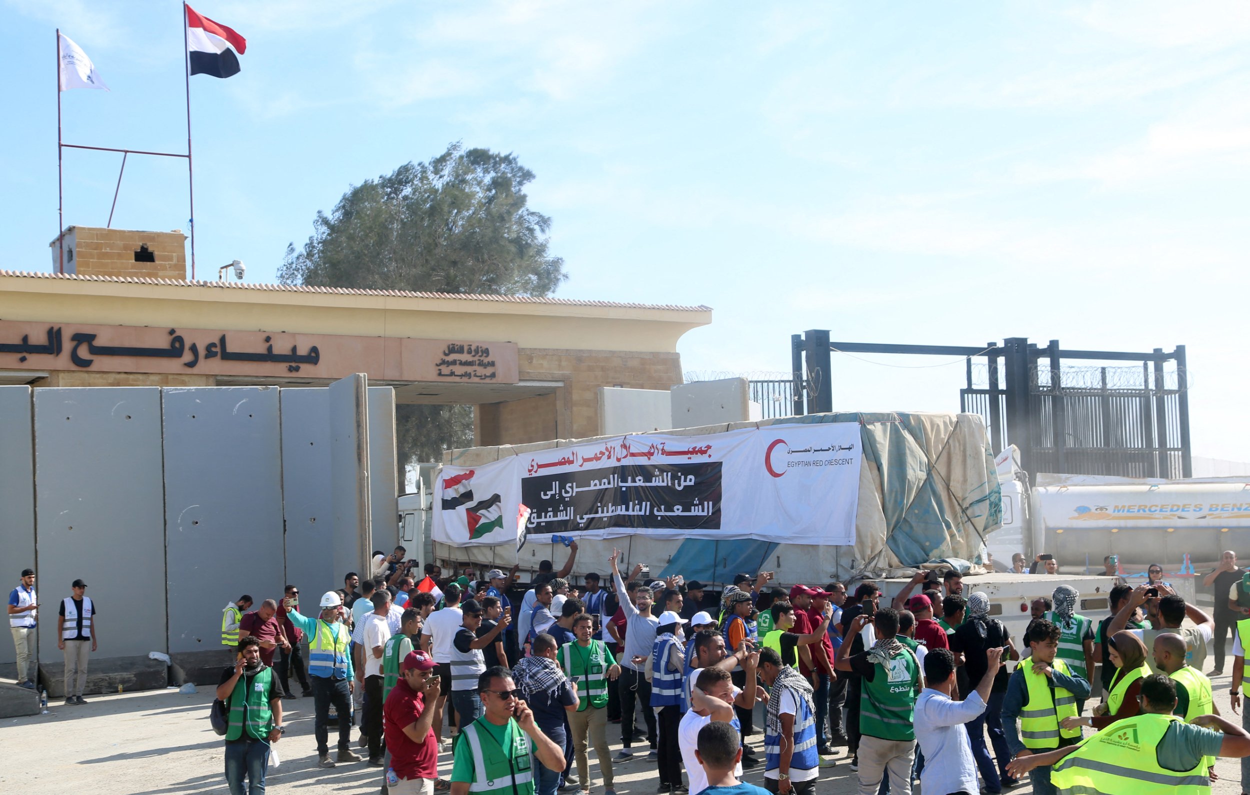 در تصویر، تعدادی از افراد در حال برگزاری یک تجمع یا اعتراض هستند. مردم در جلو ورودی یک ساختمان بزرگ ایستاده‌اند و برخی از آنها جلیقه‌های شرایطی به تن دارند. در پس‌زمینه، بنری بزرگ با متنی به زبان عربی و تصاویری از علم‌ها به چشم می‌خورد. همچنین در بالای این ساختمان، علم‌های کشورها در اهتزاز هستند. فضای تجمع به نظر شلوغ و پر انرژی می‌رسد.