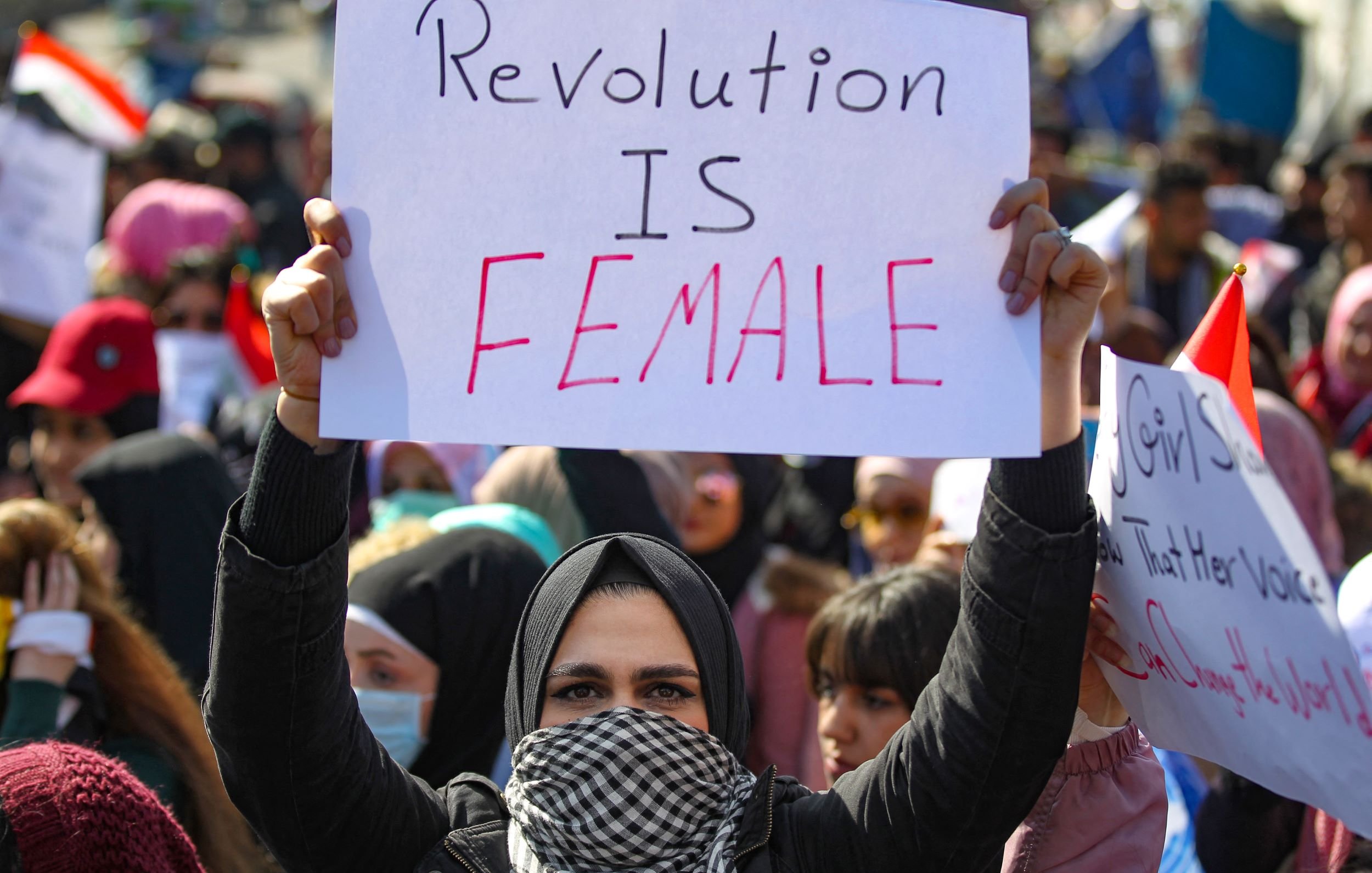L'image montre une manifestation où des personnes, en majorité des femmes, se rassemblent pour revendiquer leurs droits. Au premier plan, une femme en hijab tient une pancarte avec l'inscription "Revolution is Female". On peut voir en arrière-plan d'autres manifestants, certains portant des drapeaux et d'autres pancartes. L'atmosphère semble animée et déterminée, symbolisant la lutte pour l'égalité et l'émancipation.