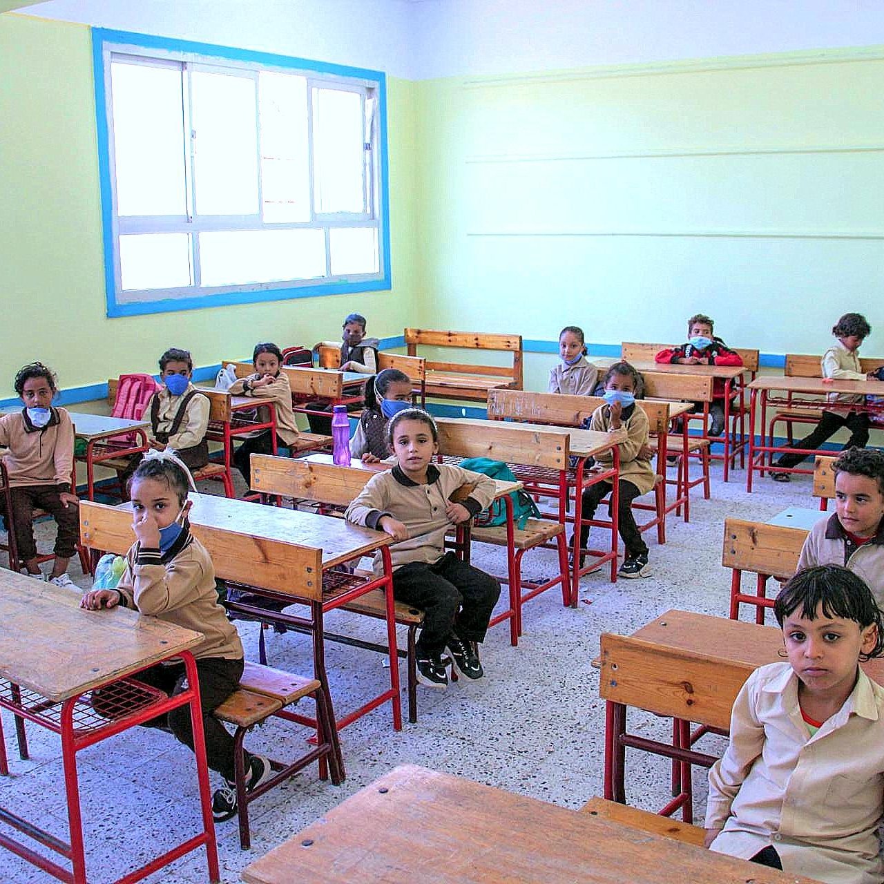 L'image montre une salle de classe avec plusieurs élèves assis à des bureaux en bois. Les enfants portent des uniformes beige et plusieurs d'entre eux ont des masques sur le visage. La pièce est lumineuse, avec des murs peints en jaune clair et des fenêtres qui laissent entrer la lumière. Les élèves semblent attentifs, certains regardant vers l'appareil photo. Les bureaux sont espacés, et il y a quelques fournitures scolaires visibles sur les tables.