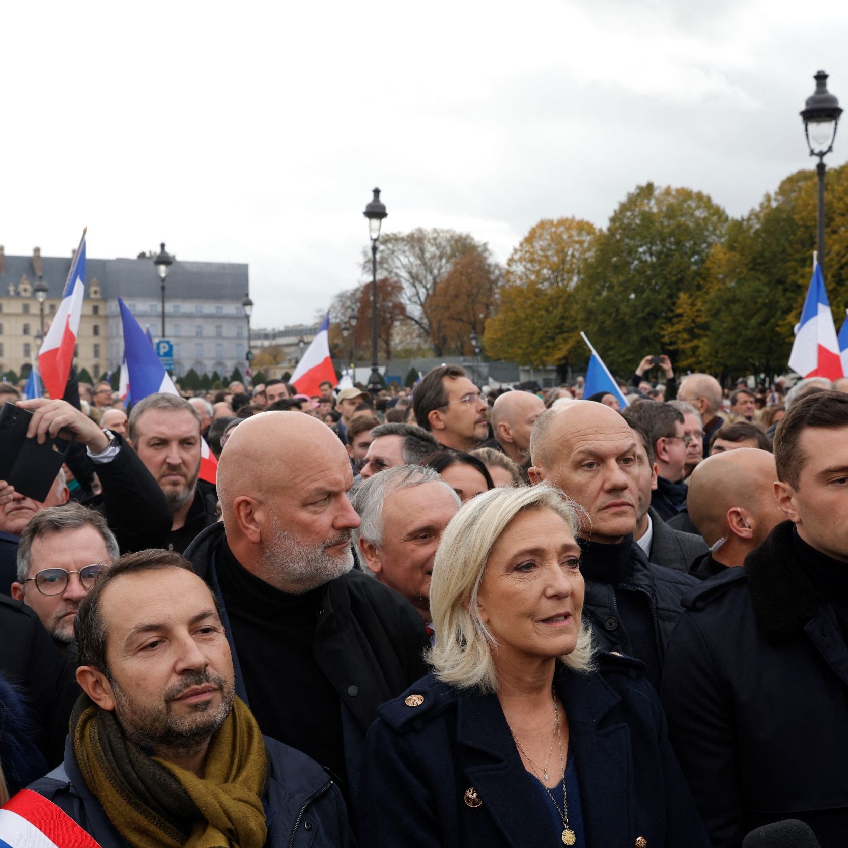 L'image montre une foule rassemblée lors d'un événement en plein air. Au premier plan, plusieurs personnes sont visibles, dont certaines portent des écharpes tricolores aux couleurs du drapeau français. On aperçoit également des drapeaux français agités par des membres de la foule. L'arrière-plan présente des bâtiments historiques et un ciel nuageux, ce qui donne une atmosphère de mobilisation. Les expressions des personnes semblent sérieuses et engagées, témoignant de l'importance de l'événement.