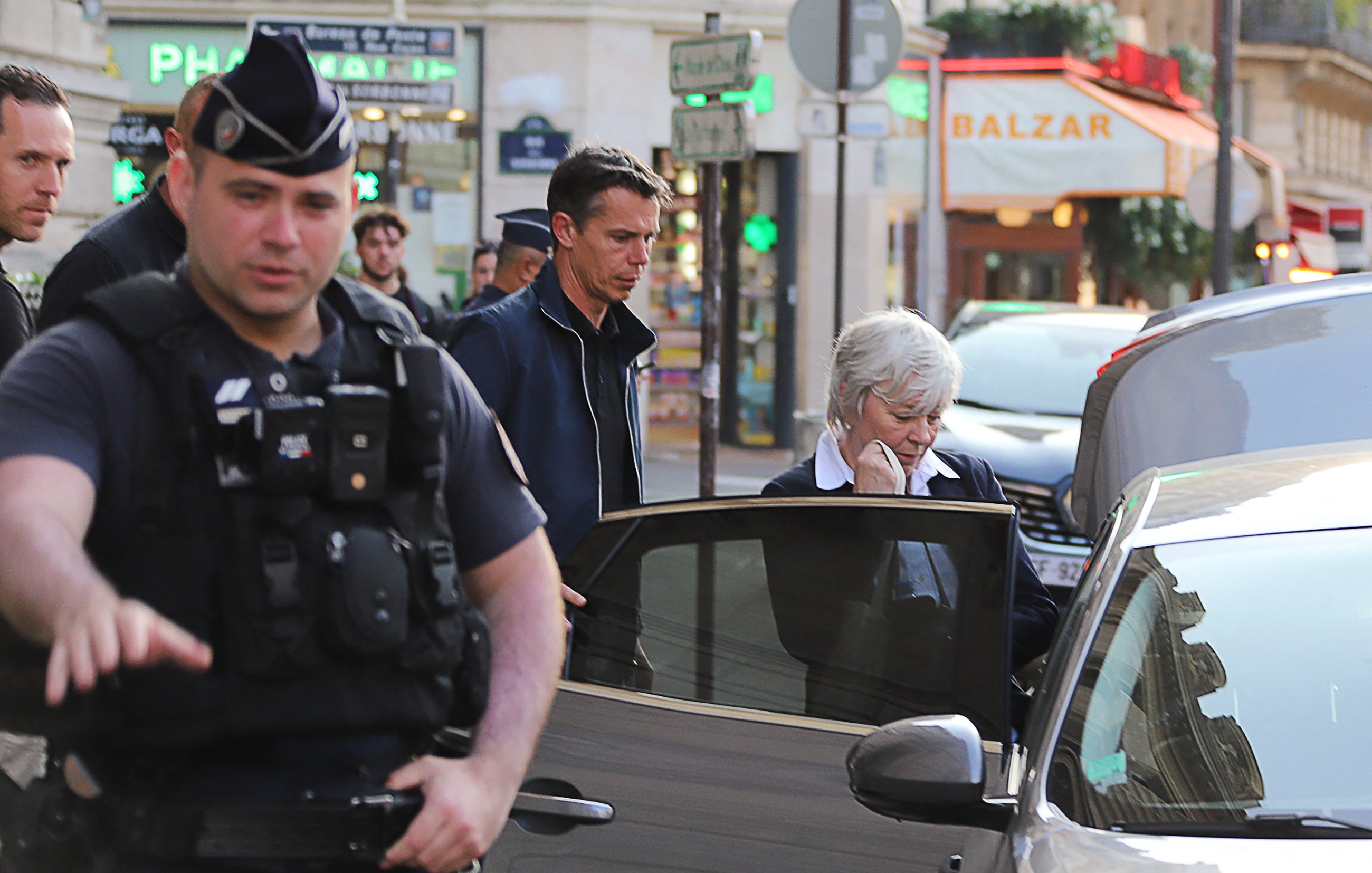 The image shows a scene on a city street. In the foreground, a police officer in uniform appears to be directing traffic or overseeing an incident. To the right, there are two individuals: a man in a dark jacket and a woman with short white hair, who seems to be getting into a car. The background features buildings and some signage, suggesting a busy urban environment. The atmosphere looks quite active, likely indicative of a recent event or situation requiring police presence.