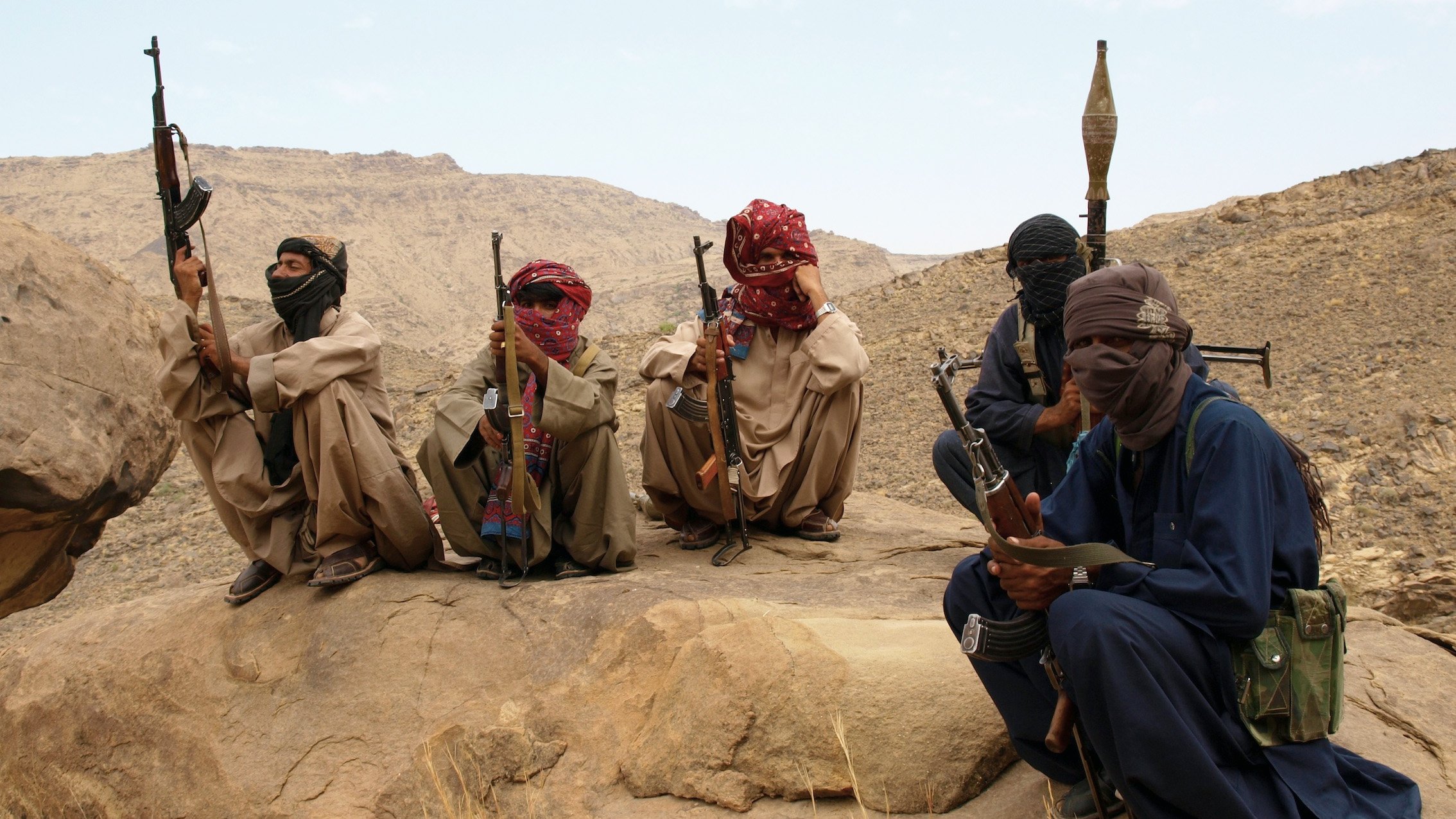 L'image montre un groupe de personnes assises sur des rochers dans un paysage désertique. Elles portent des vêtements traditionnels, masquent partiellement leur visage avec des foulards et tiennent des armes. L'environnement est montagneux et aride, avec des formations rocheuses en arrière-plan. L'ambiance semble sérieuse et reflète une situation de tension.