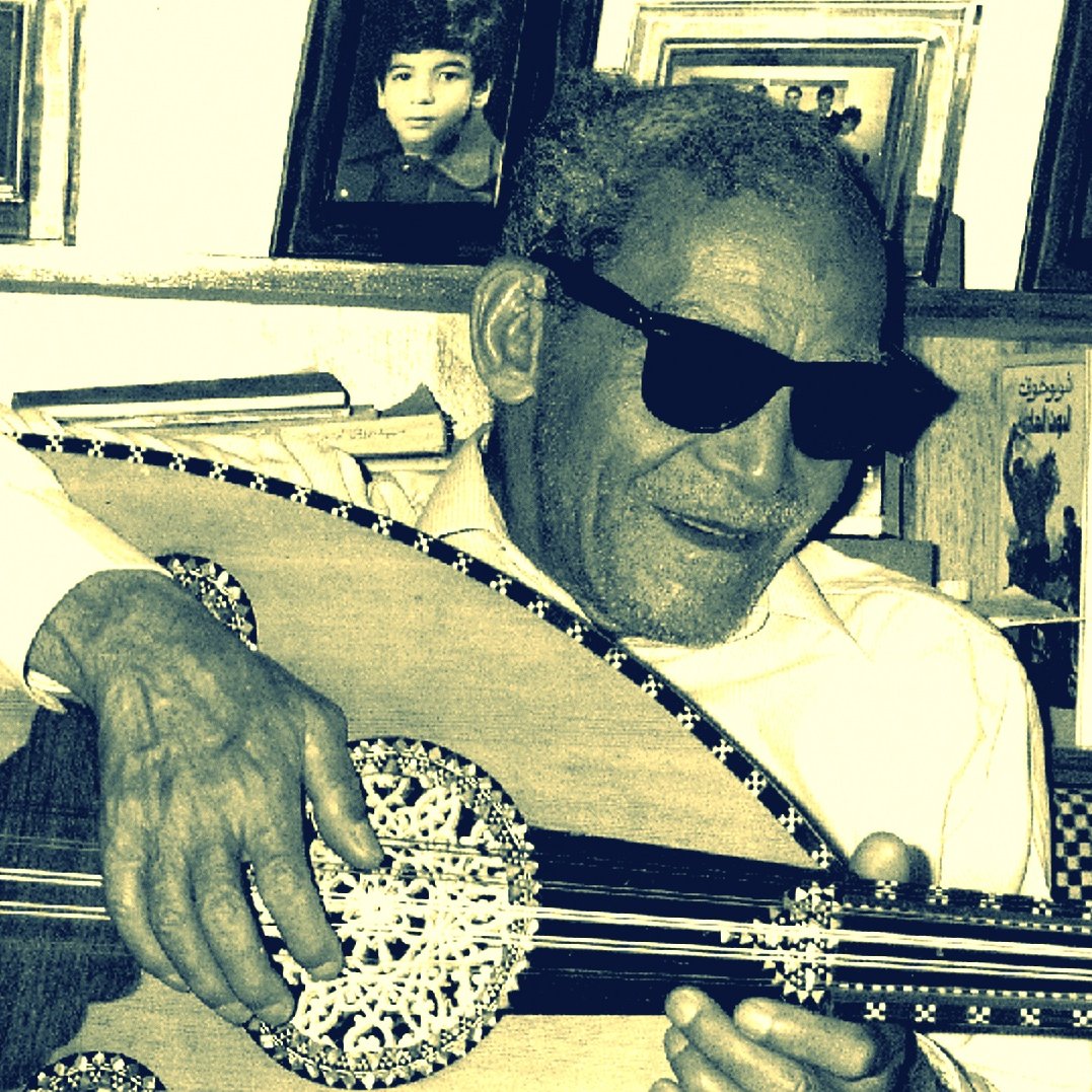 L'image montre un homme âgé portant des lunettes de soleil, assis et jouant de l'oud, un instrument de musique traditionnel. Il semble concentré sur sa musique, avec un sourire léger. En arrière-plan, on aperçoit des photos encadrées accrochées au mur, ce qui pourrait suggérer un environnement familial ou personnel. L'éclairage donne une ambiance nostalgique à la scène.