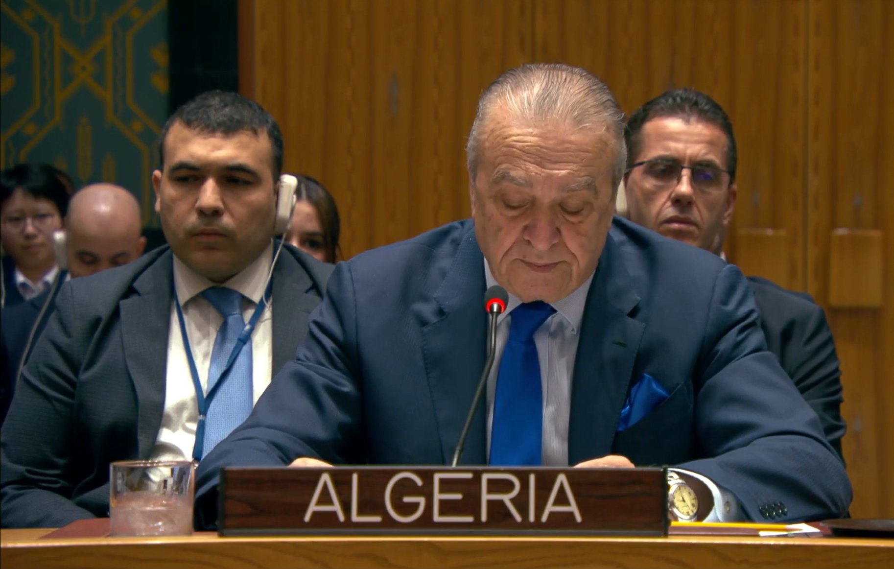 A man in a suit speaks at a podium labeled "Algeria" in a formal setting with delegates behind him.
