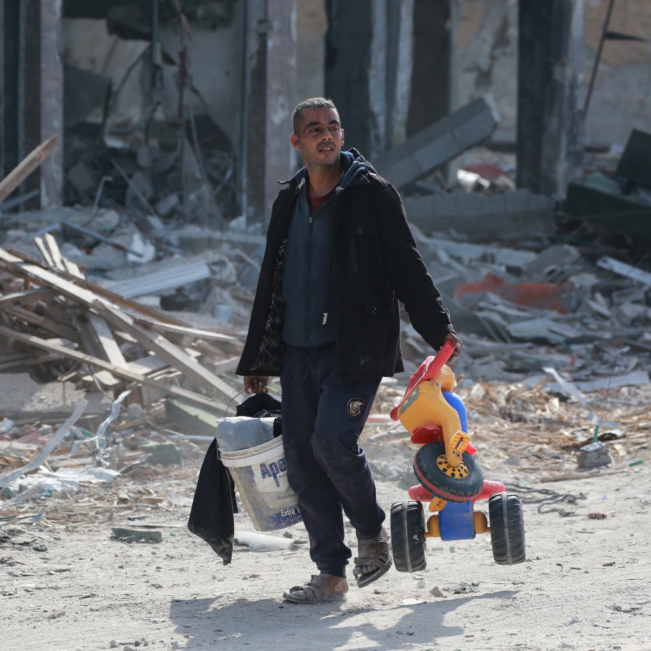 L'image montre un homme marchant dans un paysage dévasté. Il porte un seau de peinture dans sa main gauche et un jouet pour enfant, un petit véhicule à roues, dans sa main droite. L'environnement autour de lui est marqué par des débris et des ruines, suggérant un endroit qui a subi des destructions importantes. L'homme a l'air déterminé malgré la situation difficile.