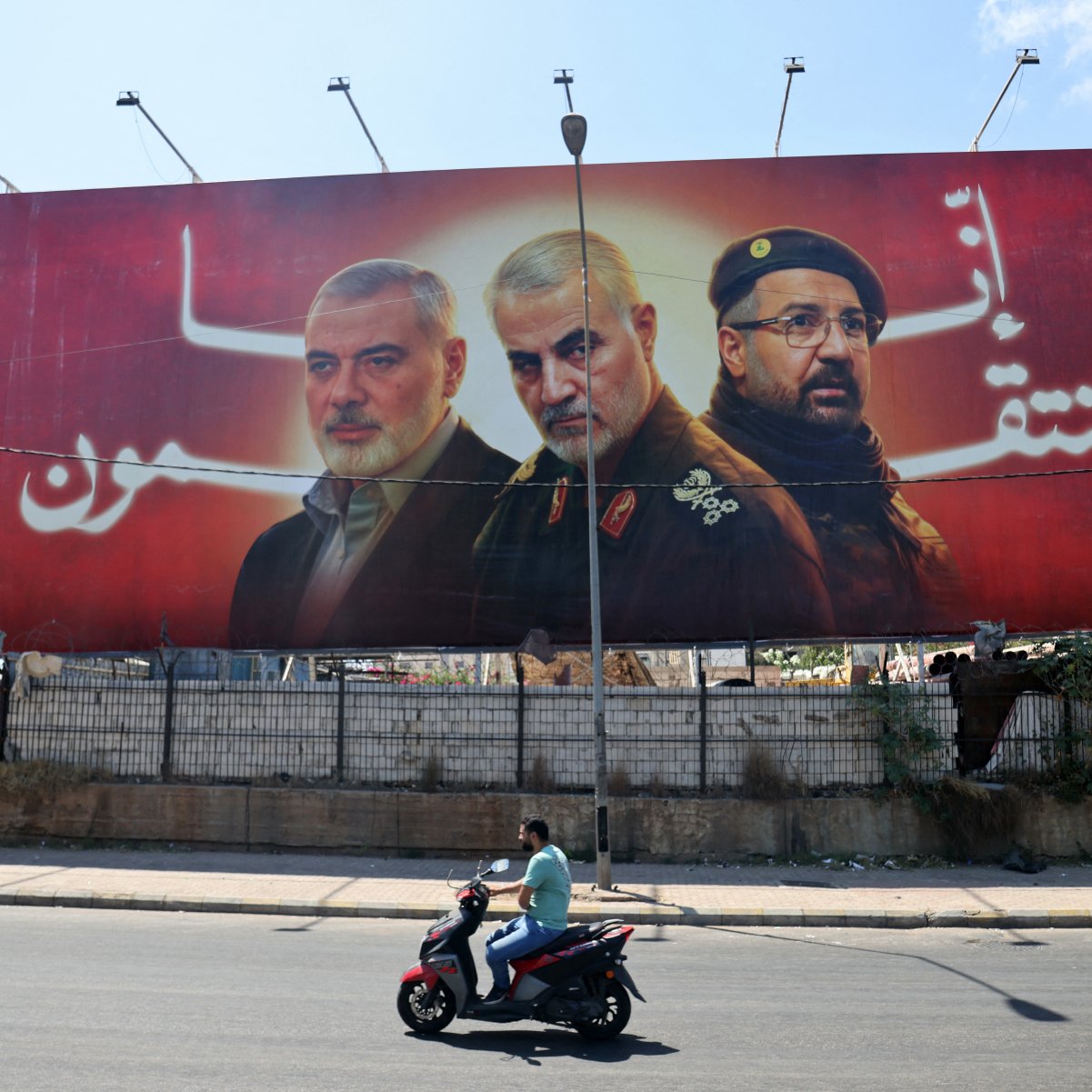 الصورة تظهر لوحة إعلانات ضخمة تحمل صور ثلاثة أشخاص. الخلفية حمراء اللون، وتبدو وكأنها تتضمن رسائل وطنية أو سياسية. في المقدمة، يوجد رجلان يواجهان الكاميرا ورجل آخر يُظهر جزءًا من وجهه، مما يوحي بأهمية شخصياتهم في السياق المعروض. في أسفل اللوحة، يُظهر شخص يقود دراجة نارية، مما يضيف حركة في الصورة ويعكس الحياة اليومية في المدينة.